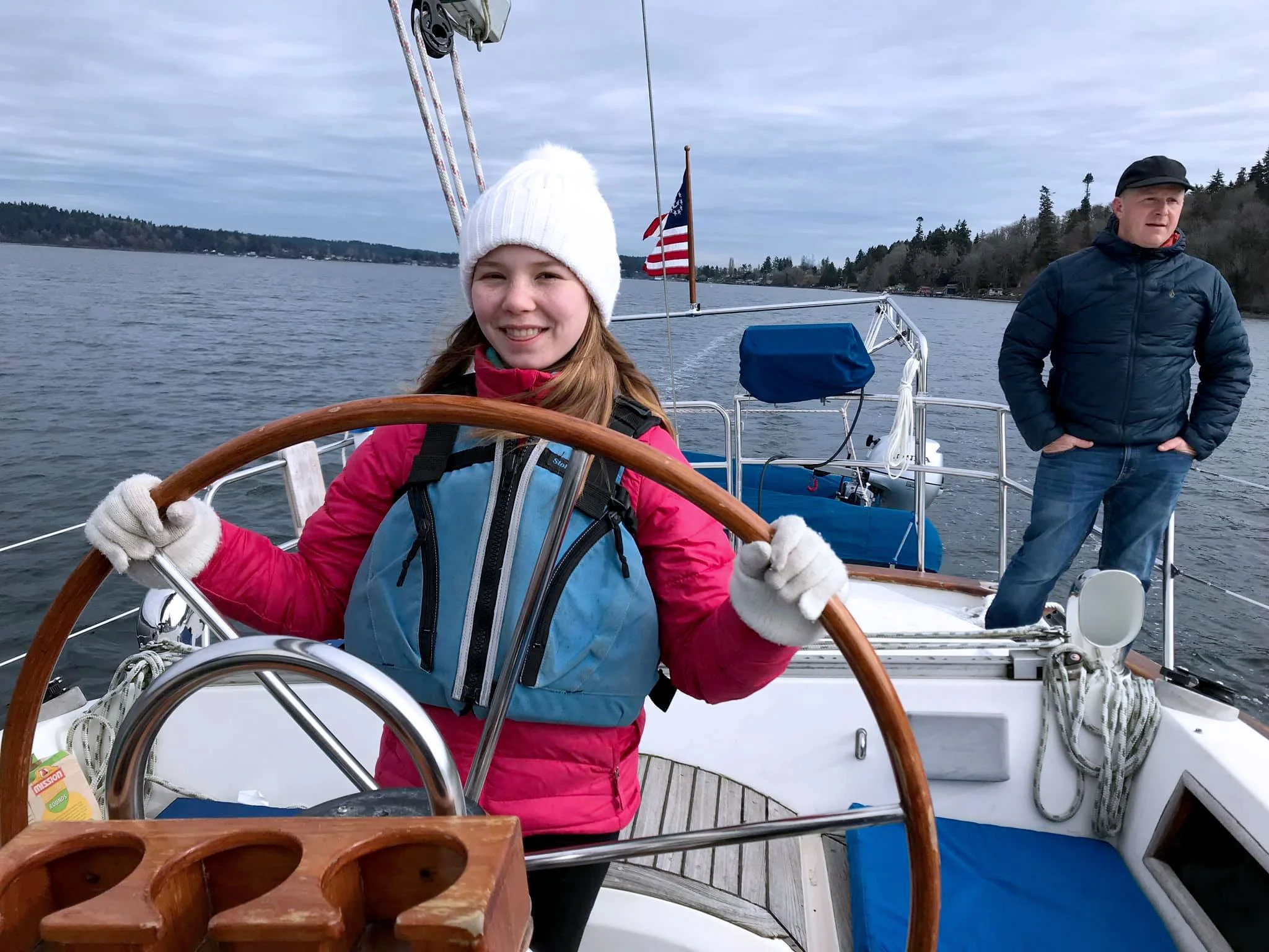 A young girl wearing a white knit hat and gloves, smiling while steering a boat on a body of water with an American flag in the background. A man stands behind her, wearing a dark jacket and cap, looking to the side. The boat has a wooden steering wheel and overcast sky.