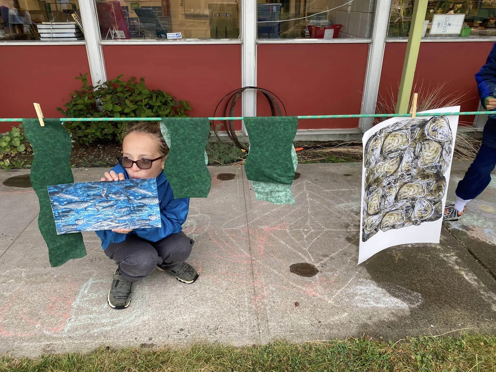 Child with sunglasses sitting on sidewalk in front of a display of seafood paintings hung on a green string, outside a storefront with large windows and red panels.