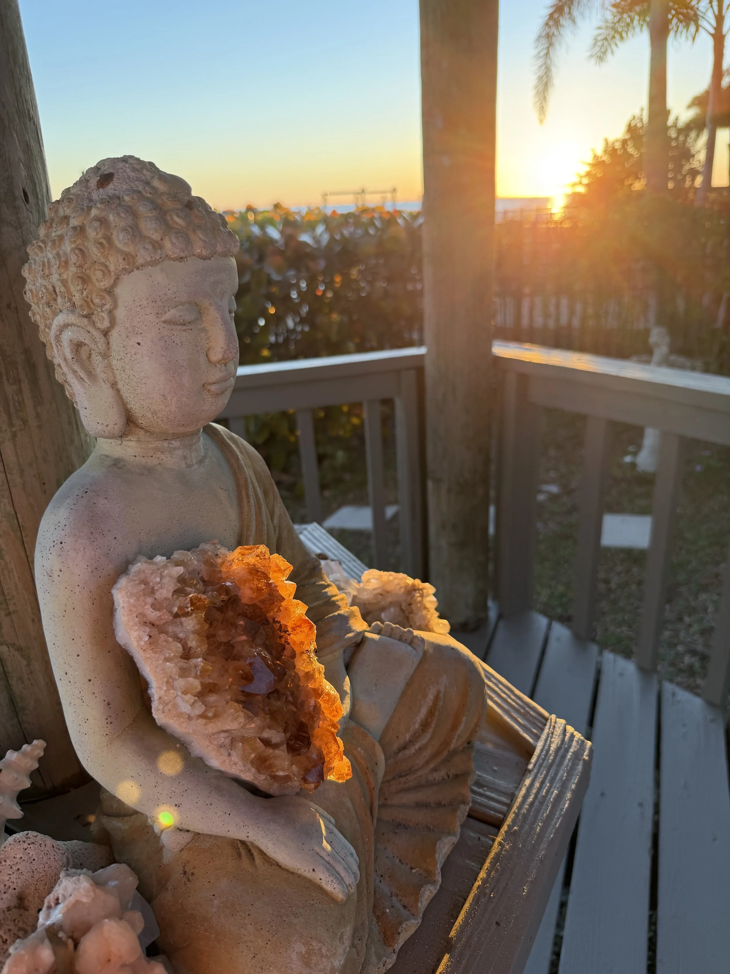 Statue of a meditative buddha figure holding a geode with crystals, with a sunset and palm trees in the background.
