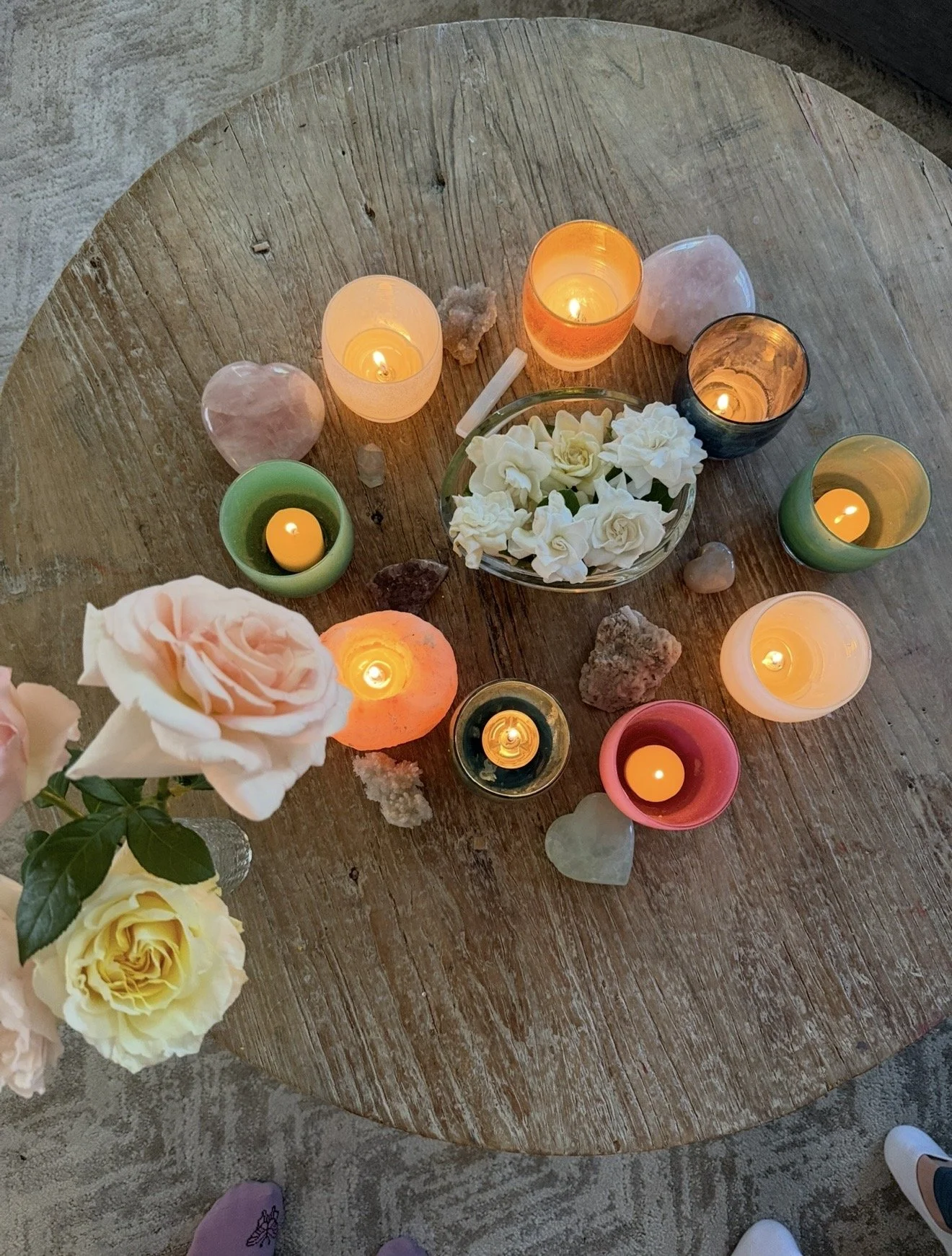 A wooden table with multiple lit candles, white roses, and various crystals arranged on it, creating a calming and cozy ambiance.