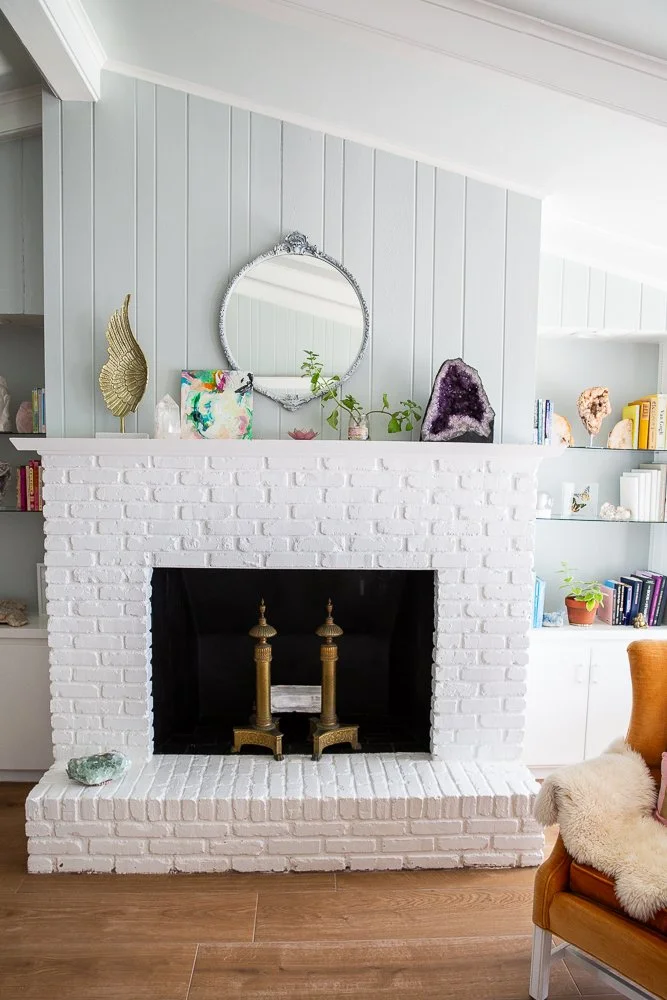 White brick fireplace with brass fireplace tools, a small mirror above, and decorative items on the mantel. To the right, part of an orange armchair with a white furry blanket. Built-in shelves with books and decor are on either side of the fireplace.
