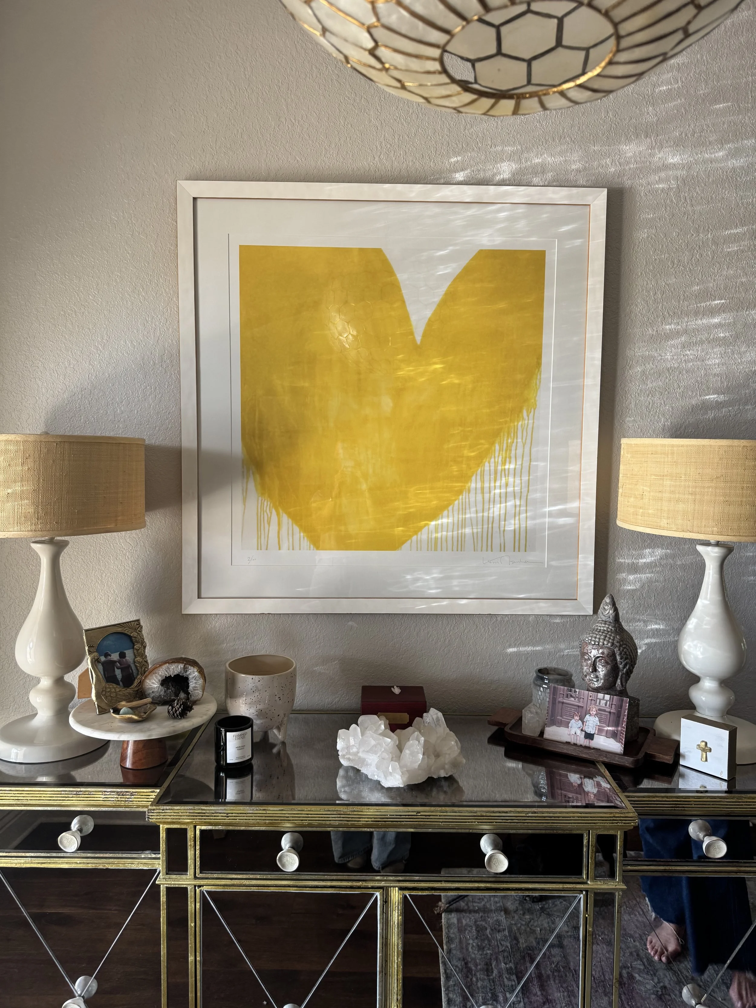 Decorative wall art featuring a large yellow heart with dripping paint at the bottom, framed and hanging on a textured wall. Below, a black table with a glass top holds various decorative items, including sculptures, candles, and framed photographs, 