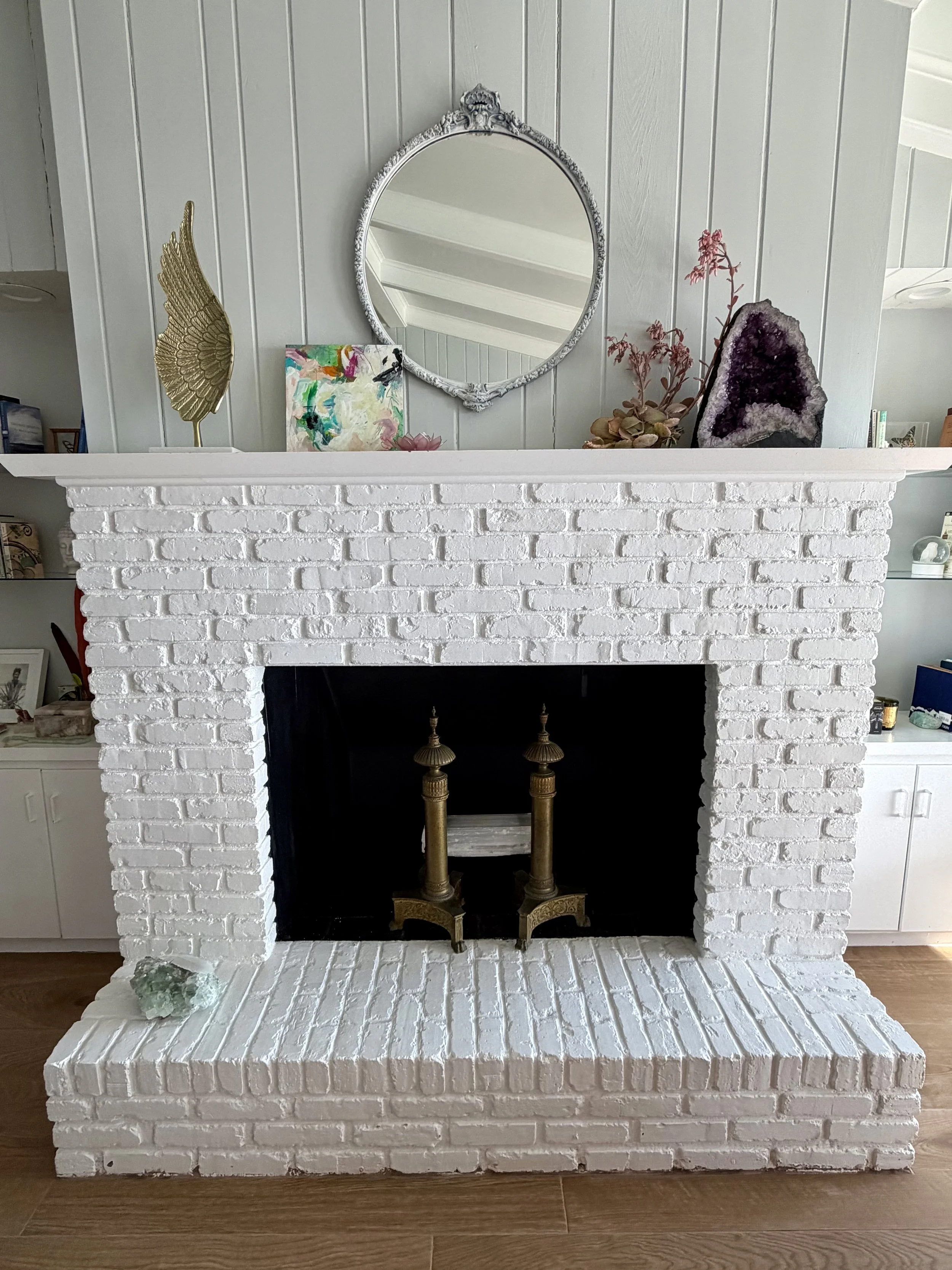 A white painted brick fireplace with decorative items on the mantel, including a framed abstract artwork, a gold wing sculpture, pink flowers, and a large purple geode. A round mirror hangs above the mantel, reflecting the ceiling and upper walls of 