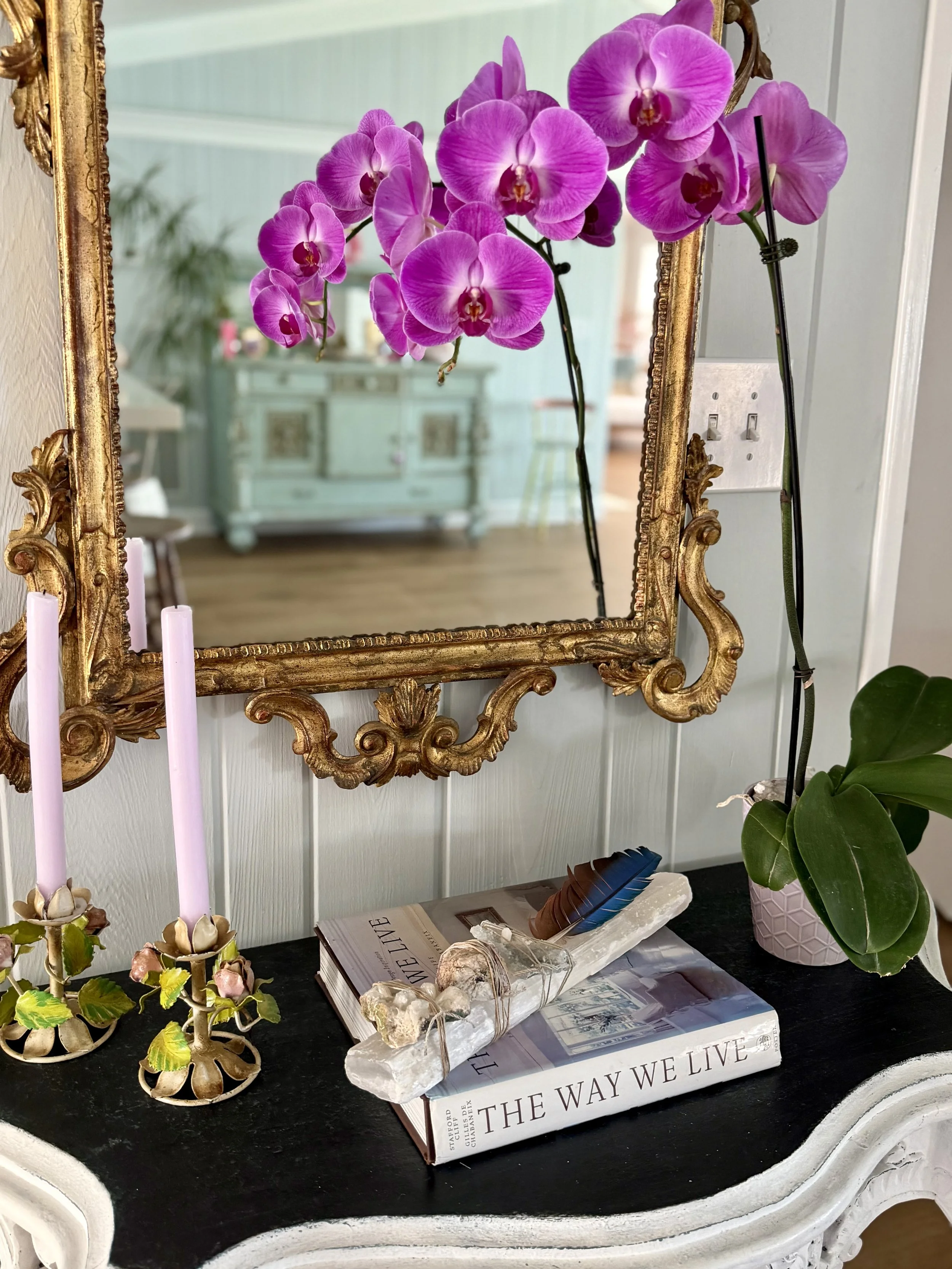 A decorative mirror with a gold ornate frame hangs above a black table. On the table, there are pink and purple orchids, a candle holder with a candle, a stack of books including one titled "THE WAY WE LIVE," and a box with decorative stones and a fe