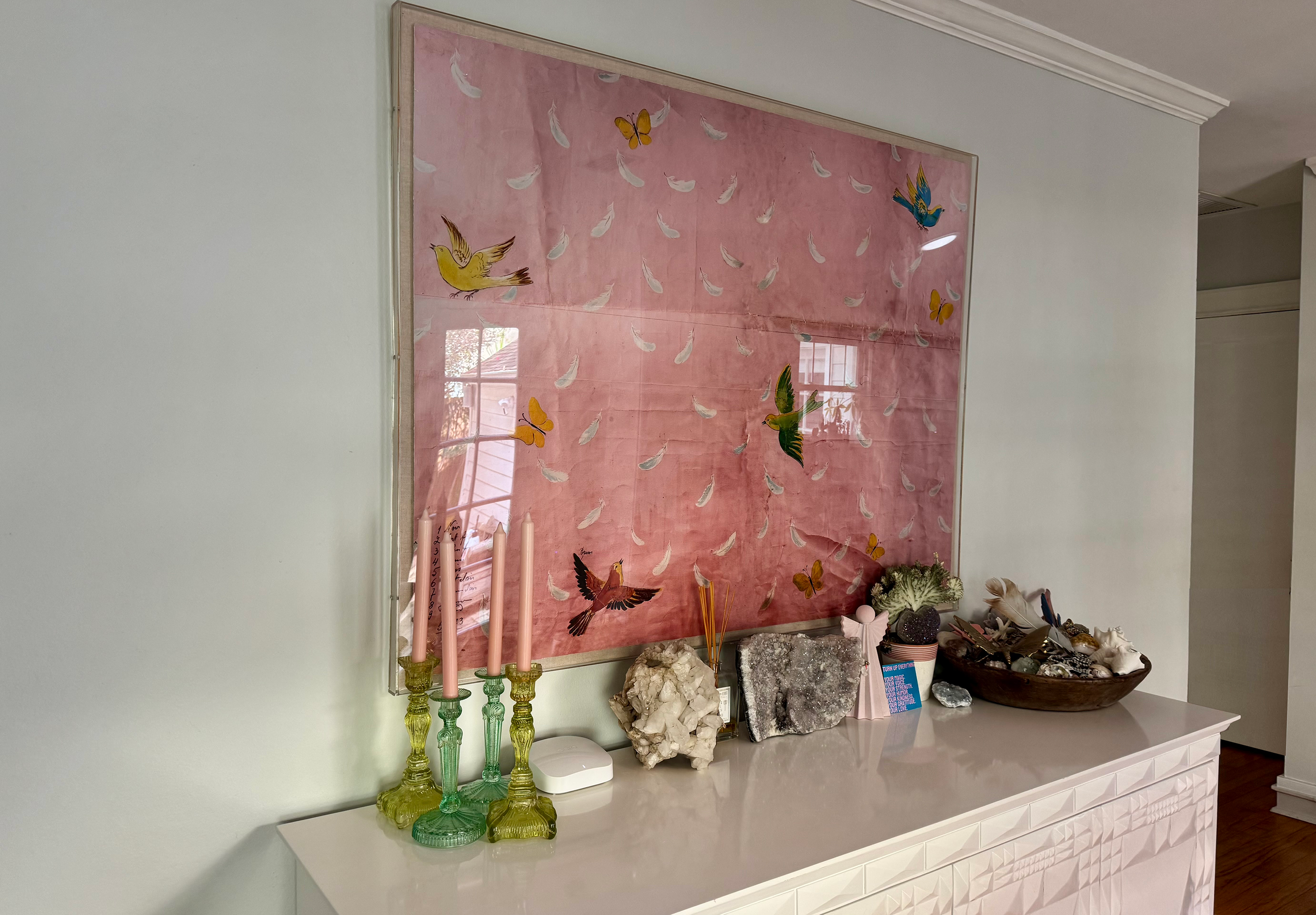 Decorative interior table display with three green glass candles, various rocks, shells, and a pink feather-themed artwork hanging on the wall behind.