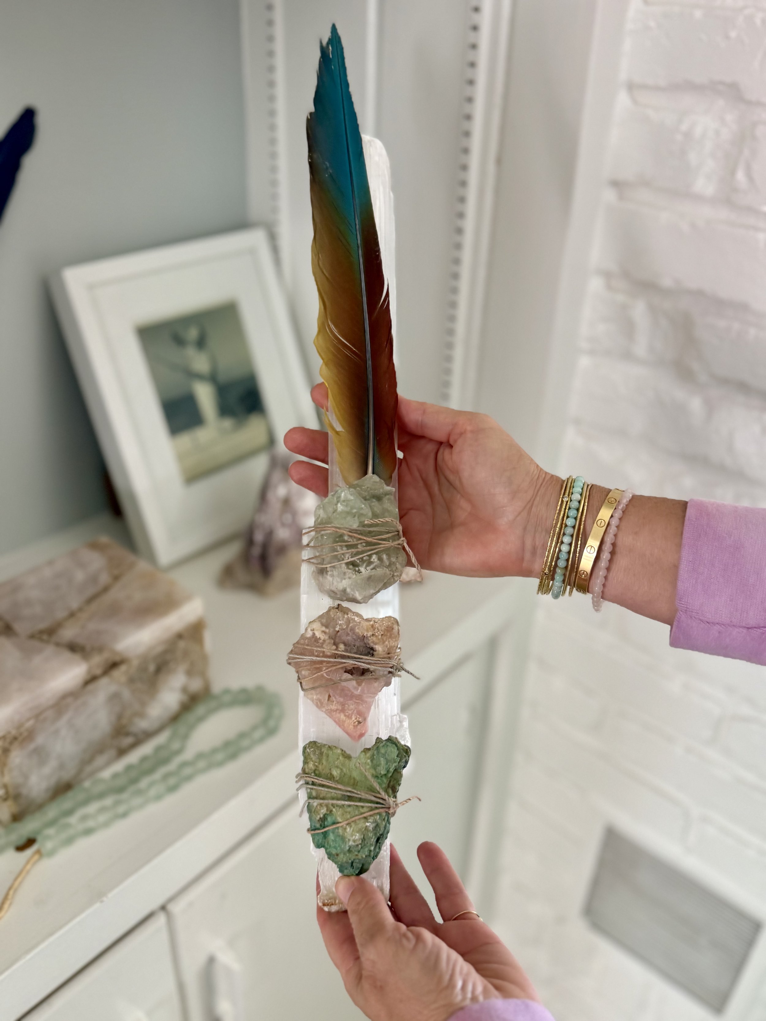 Hand-held crystal chakra healing wand with multiple semi-precious stones stacked along its length, held by a person wearing colorful bracelets, in a room with white walls and a small mirror in the background.