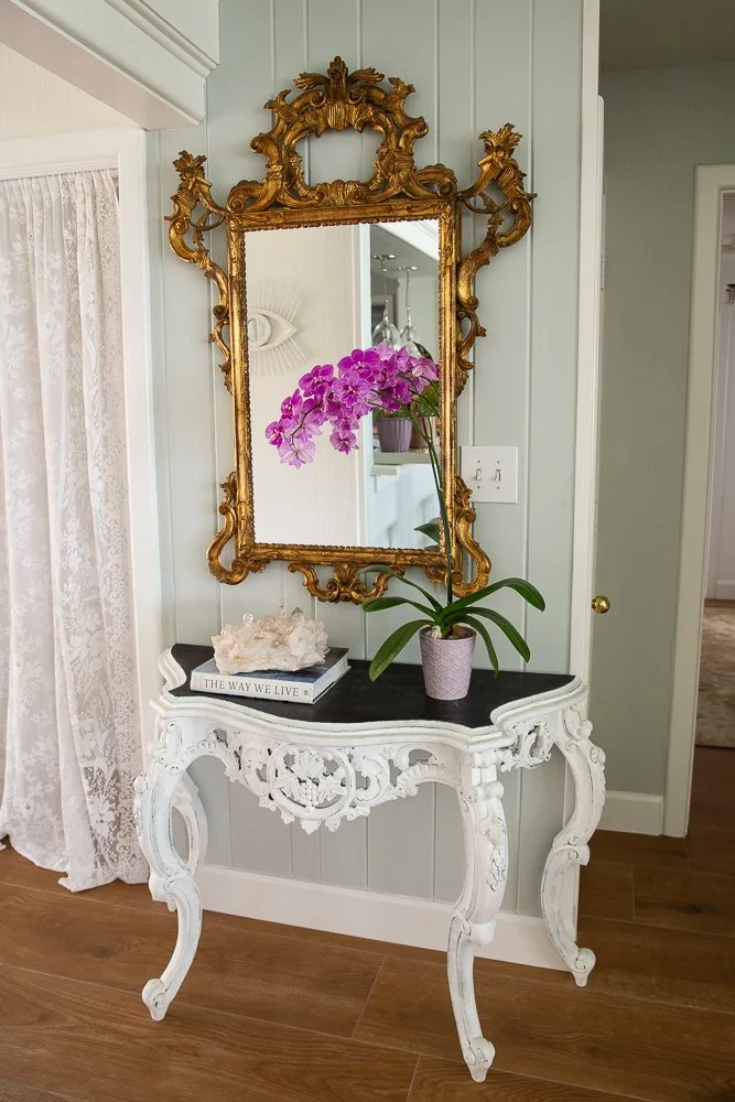 A decorative mirror with a gold ornate frame hangs above a black table. On the table, there are pink and purple orchids, a candle holder with a candle, a stack of books including one titled "THE WAY WE LIVE," and a box with decorative stones and a fe
