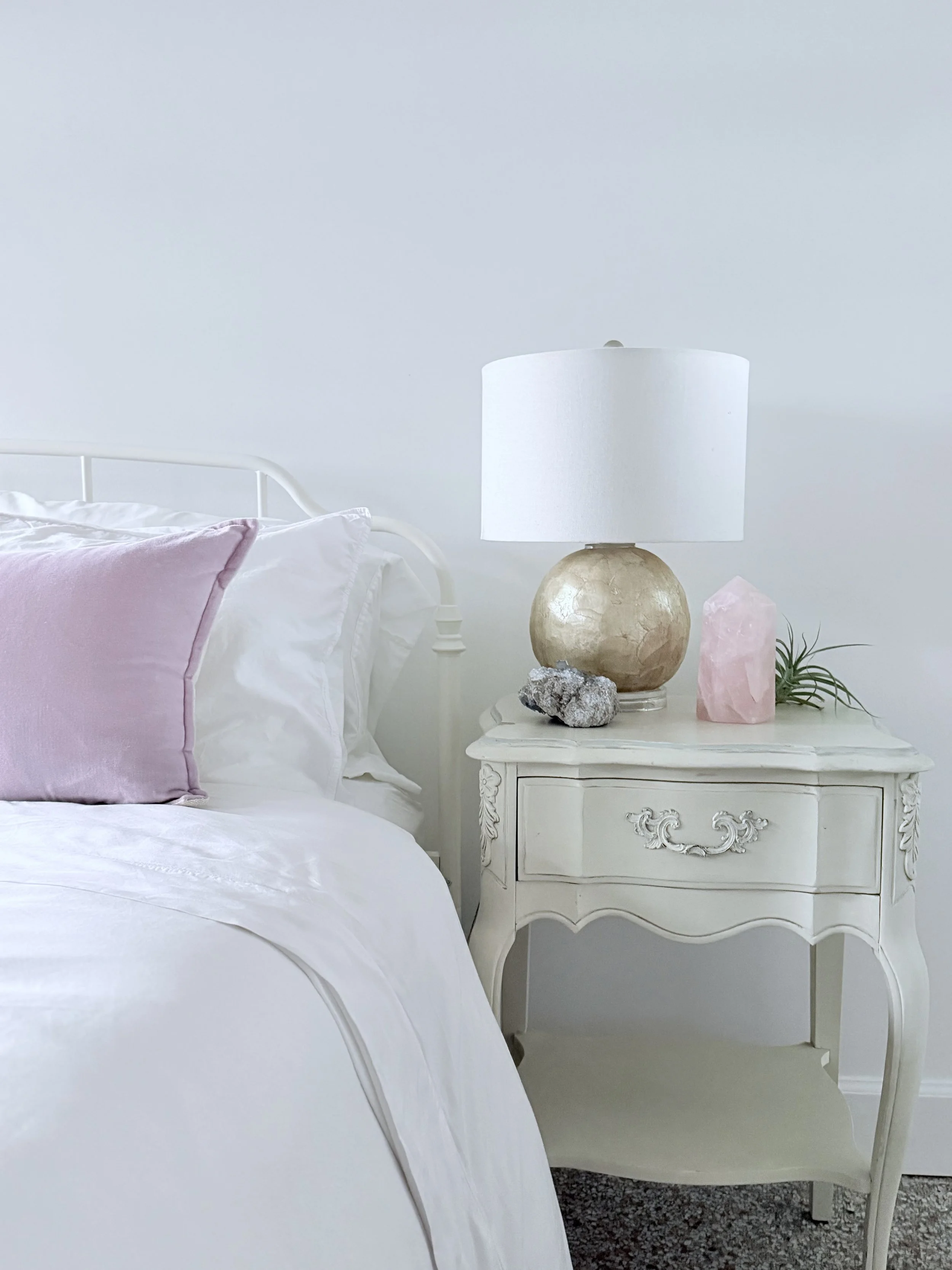 A white bed with a pink pillow and white sheets, beige nightstand with a white lamp, pink crystal, and a rock, against a white wall.