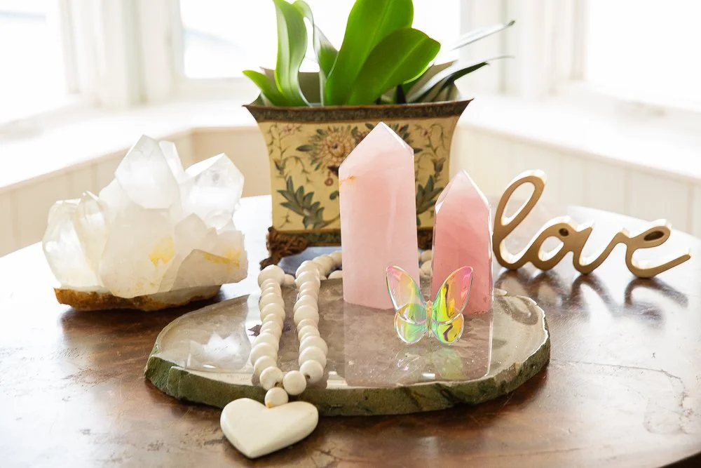 Decorative arrangement on a wooden surface with pink crystals, a white beaded necklace with a heart pendant, yellow butterfly accent, a "love" sign, a crystal cluster, and a potted plant in the background.