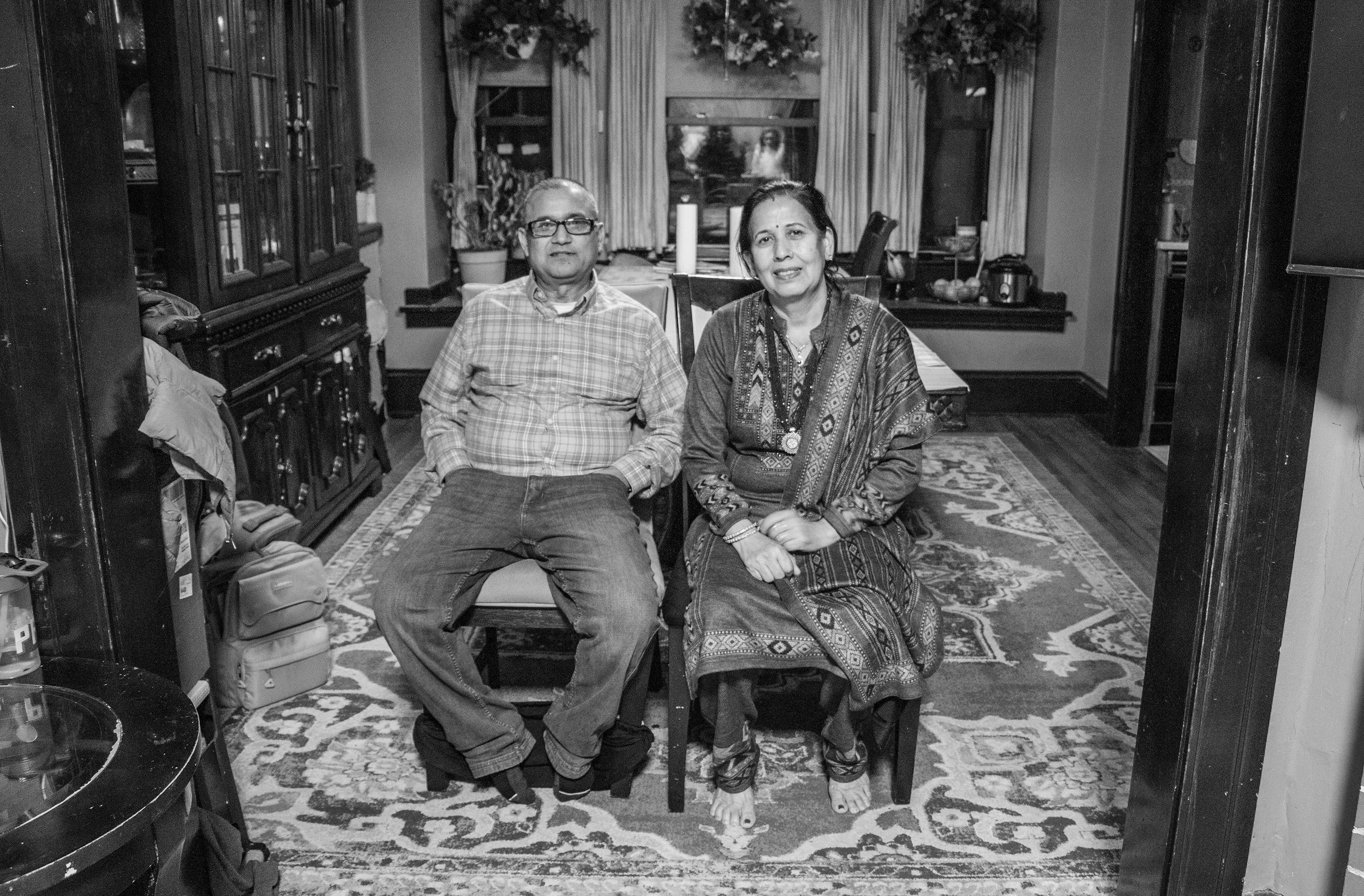 A black and white photo of an elderly man and woman sitting in a dining room. The man is wearing glasses, a checkered shirt, and jeans, while the woman is dressed in traditional attire with a patterned shawl. They are seated on chairs in front of a window with curtains and potted plants.