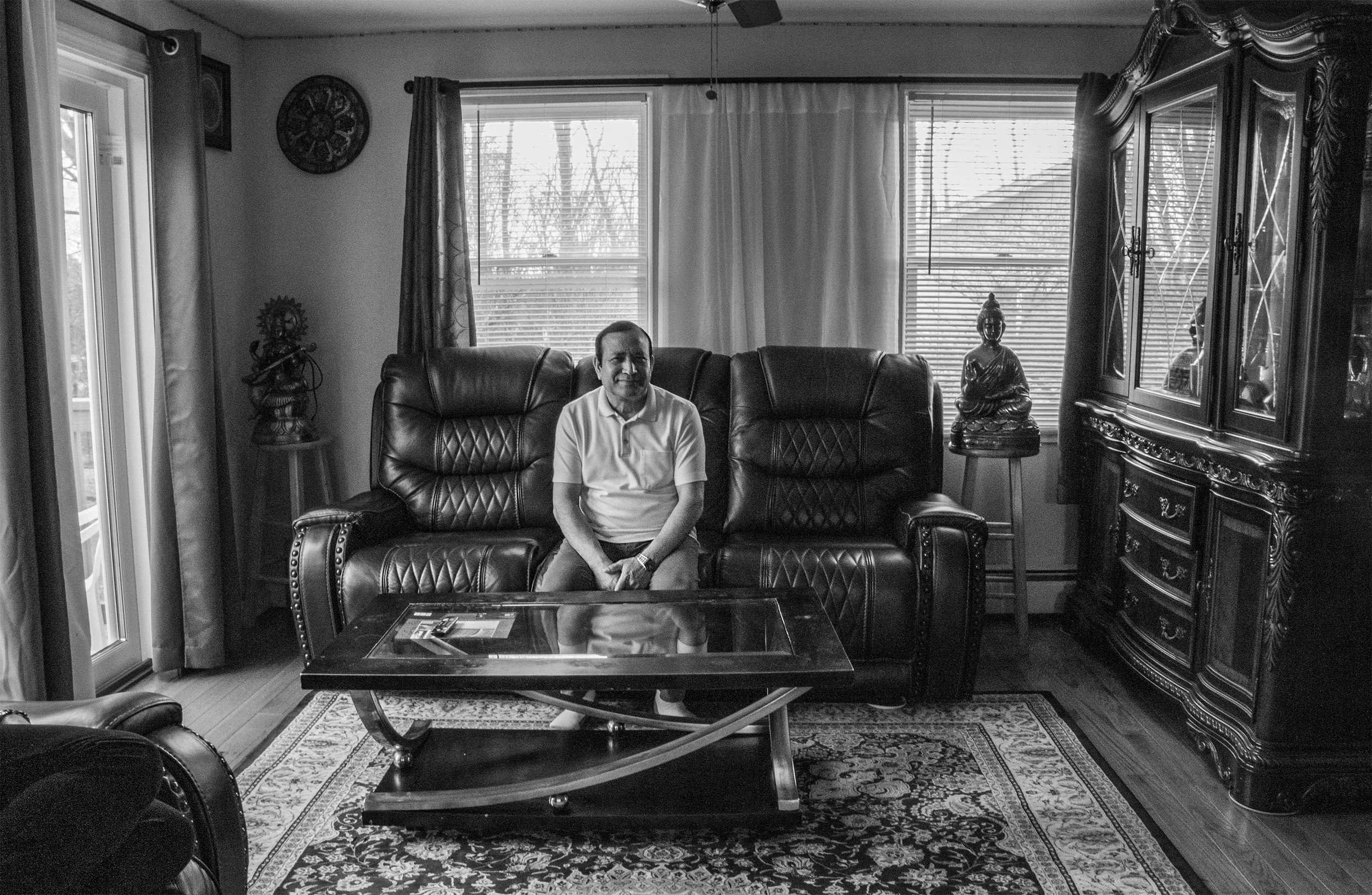 A man sitting on a black leather sofa in a living room with traditional Asian decor, including statues and a wooden cabinet, with windows and curtains in the background.