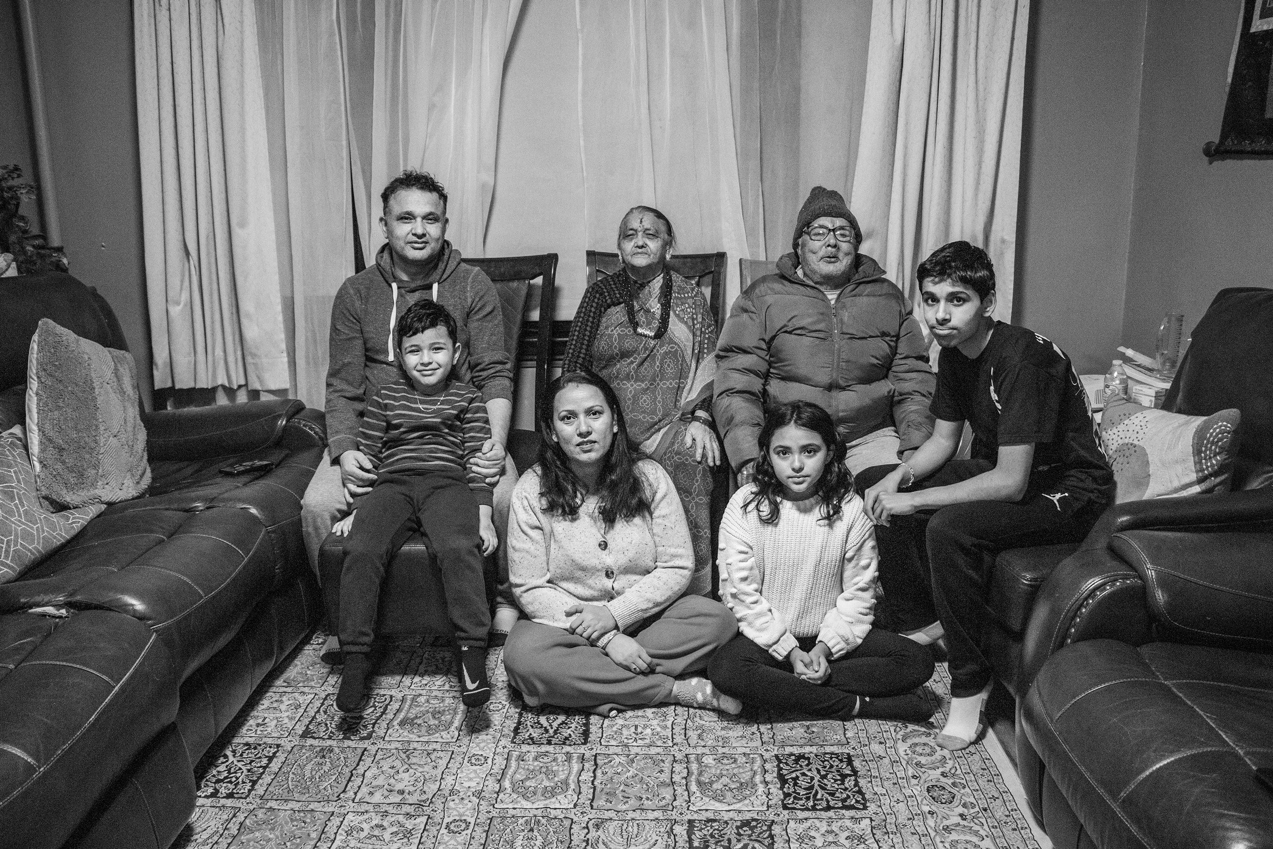A black and white photo of a large family gathered in a living room. They are seated and kneeling on the floor in front of a curtain. The group includes three older adults and five children, with sofas and decorative pillows visible in the background.