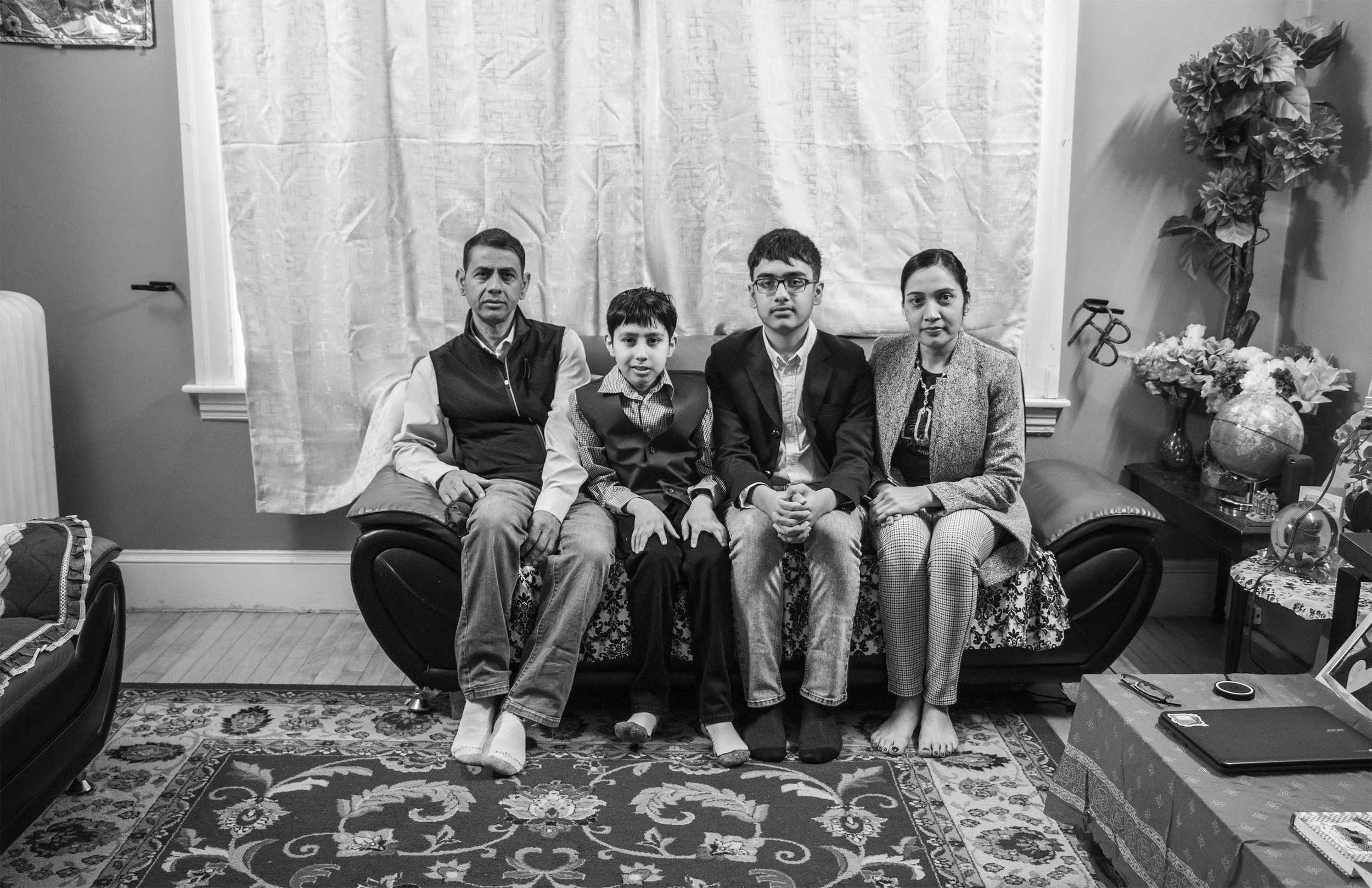 A family of five sitting on a couch in a living room, with a table in front. The room has a patterned rug, floral arrangements, and closed curtains behind them.
