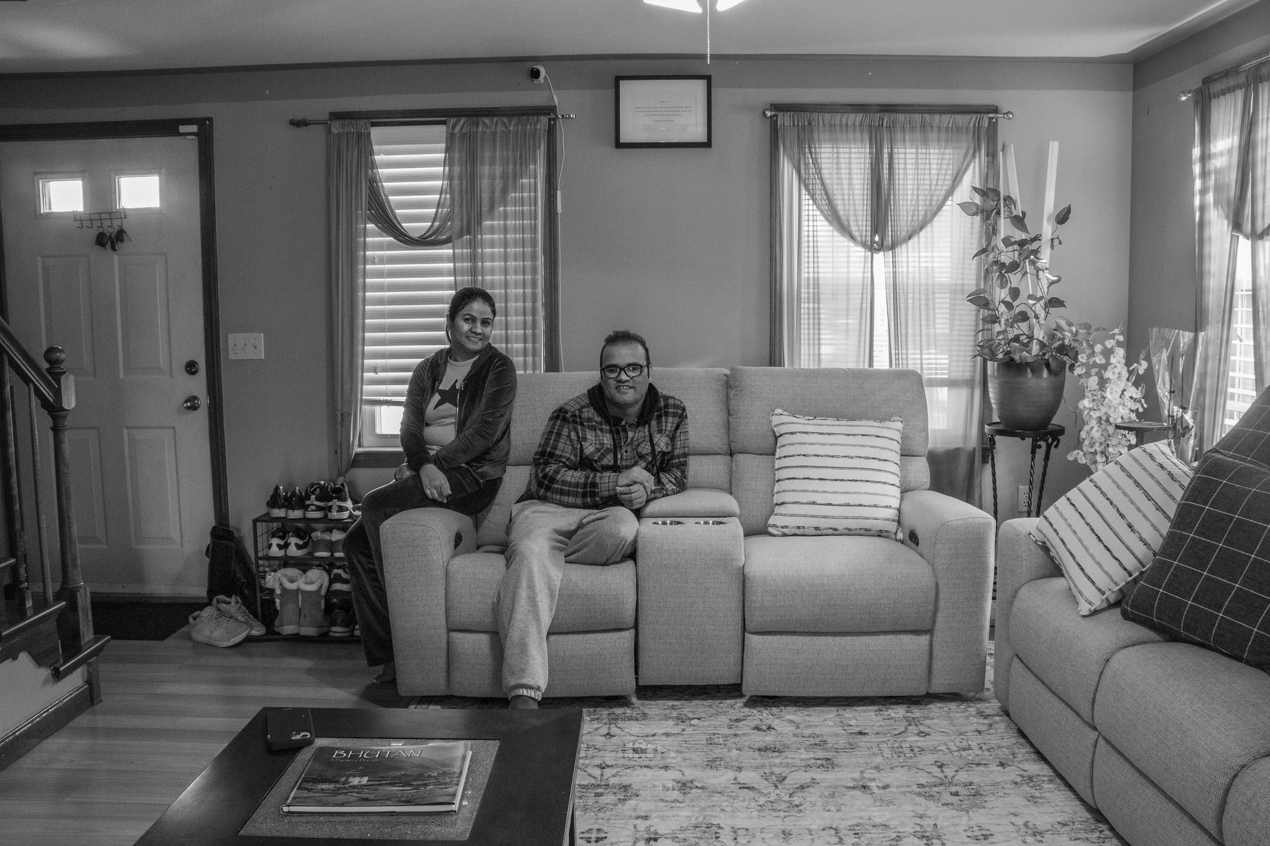 A living room with two people sitting on a couch, one woman and one man. The woman is on the left, smiling, wearing a jacket and a t-shirt with a star. The man on the right is wearing glasses and a plaid shirt, smiling, with a remote control in the middle of the couch. There are windows with curtains behind them, a plant on the right side, and a door on the left. A coffee table with magazines is in front of them.