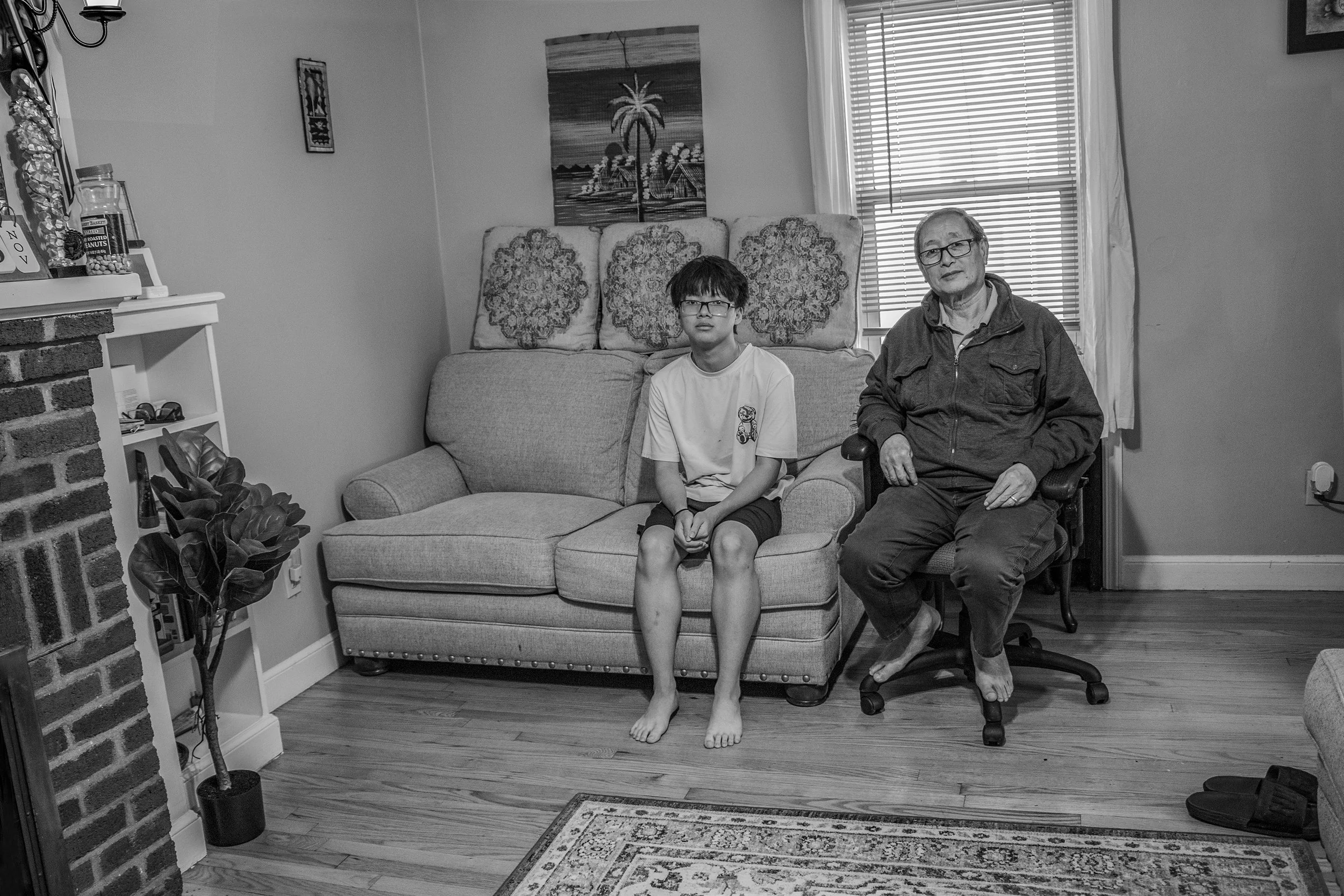 A black and white photo of a boy and an elderly man sitting on a sofa in a living room. The boy is barefoot, looking at the camera, and wearing glasses and a T-shirt. The elderly man is sitting on a rolling office chair, also barefoot, wearing glasses, and a zip-up jacket.