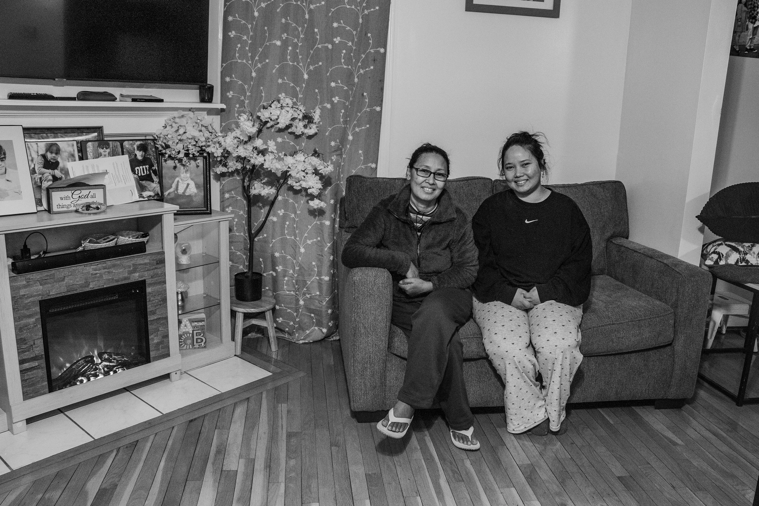 A woman and a girl sitting on a sofa in a living room, smiling, with a fireplace, photo frames, curtains, and decorative items in the background.