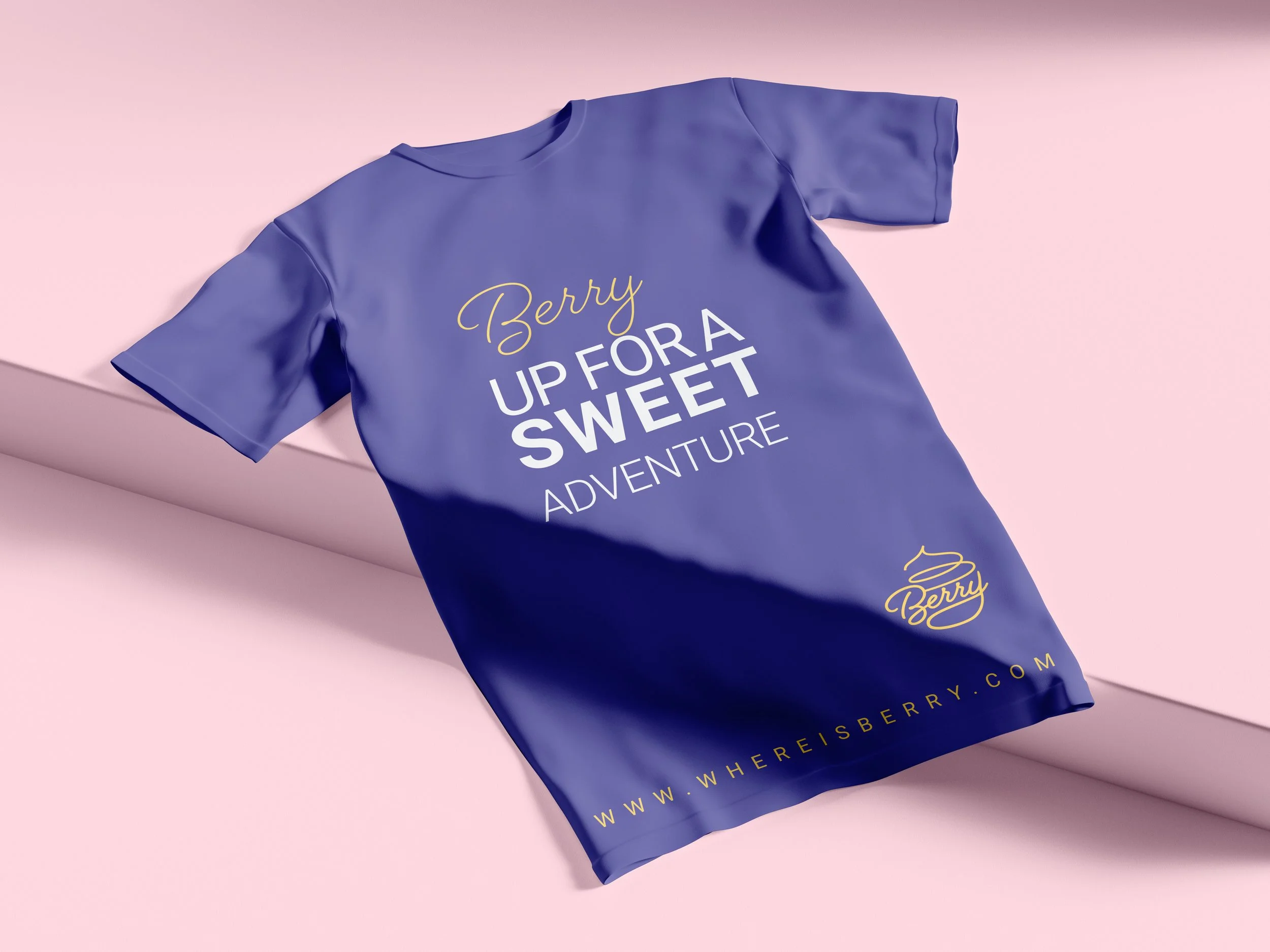 Blue T-shirt with yellow and white text that reads "Berry Up for a Sweet Adventure" and includes a small yellow logo and website URL at the bottom, laid on a pink surface.