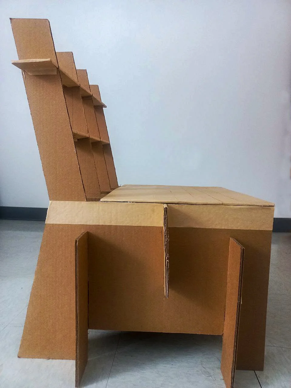 A chair made from cardboard with a high backrest, armrests, and a seat, crafted from flat pieces of cardboard assembled into a structured design.
