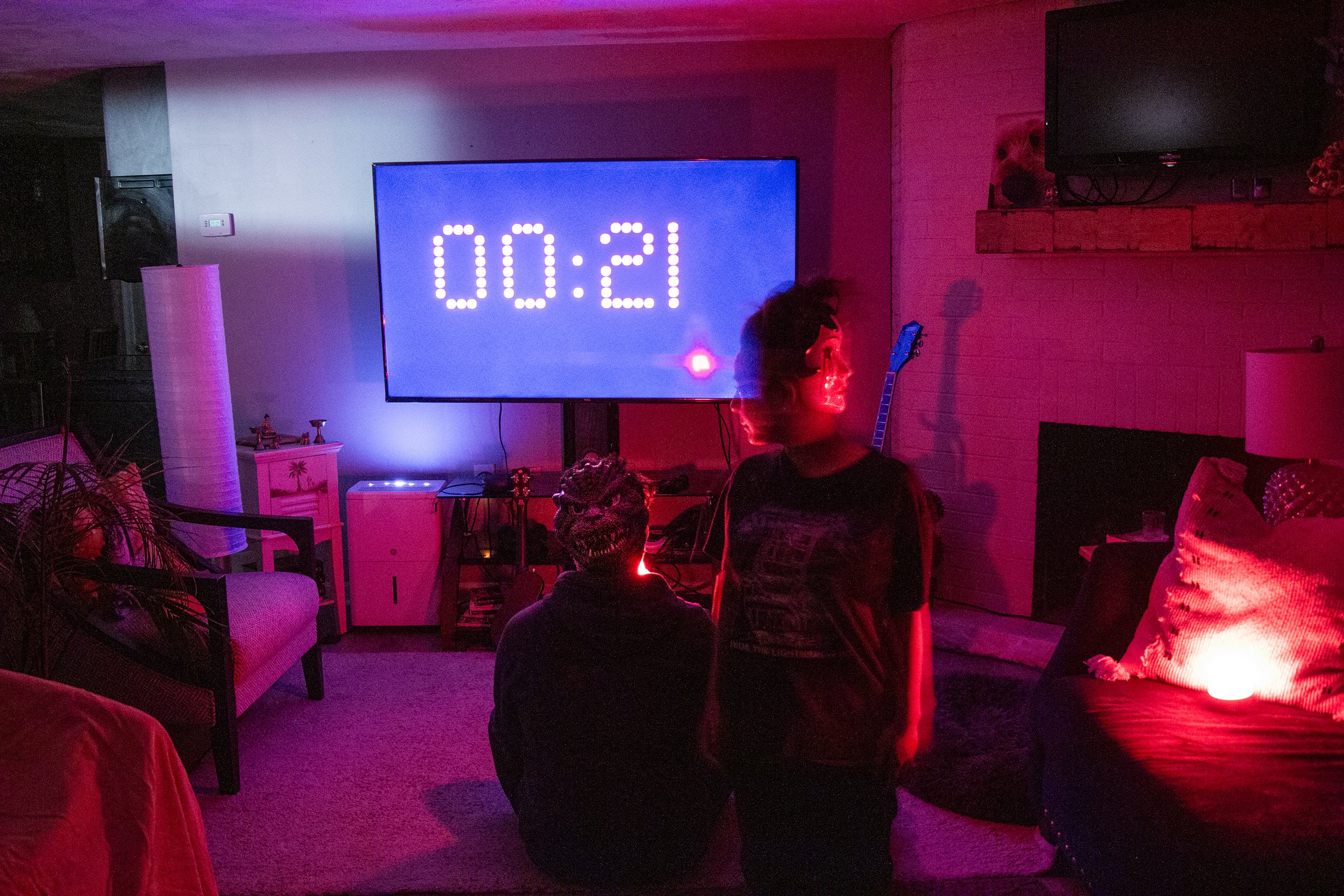 A living room illuminated with red and purple lights, with a large television screen displaying a timer showing 21:00, two people are present: one sitting on the floor wearing a mask, and the other with a messy hairstyle standing nearby, a guitar lea