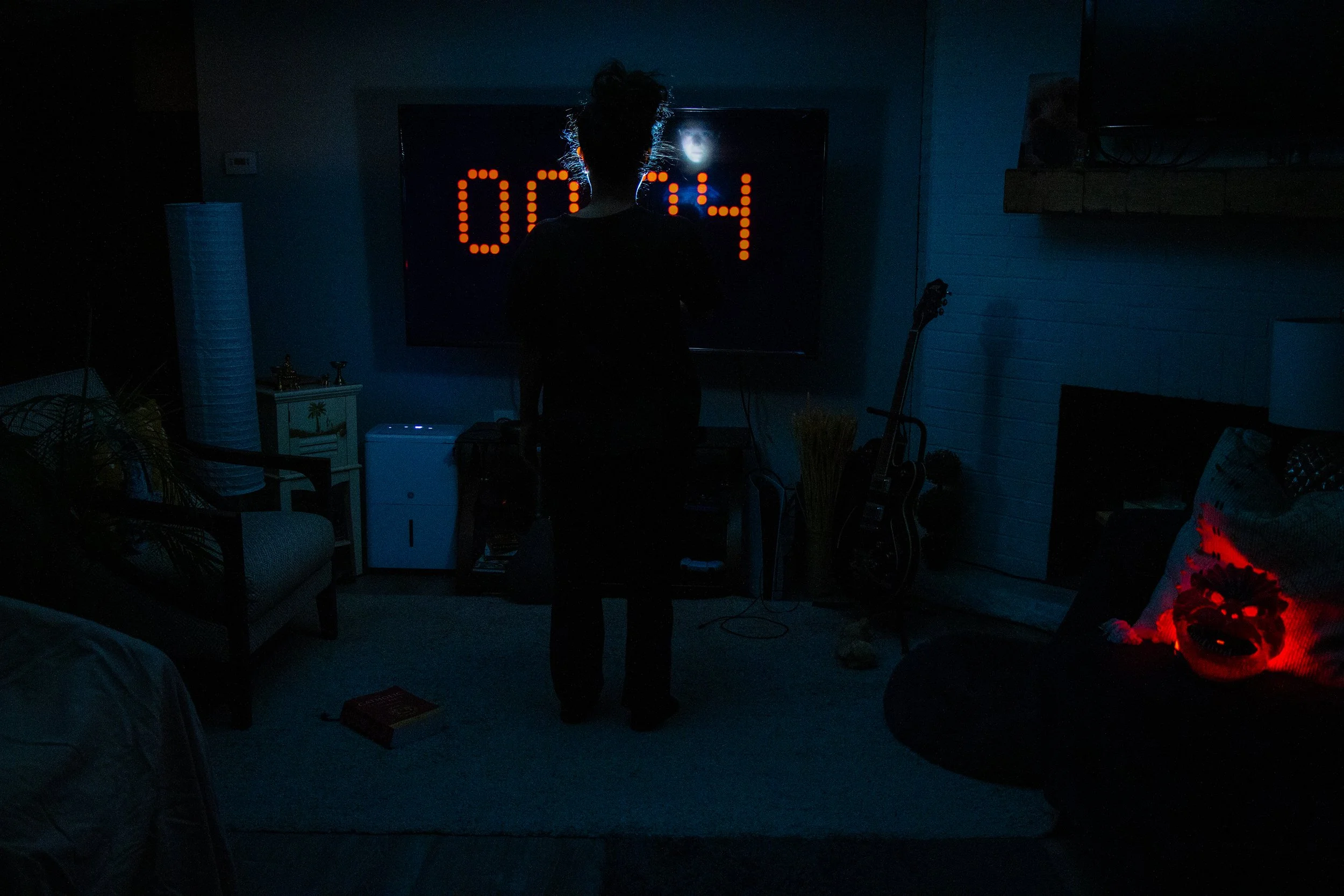 A person stands in a dark room in front of a large digital clock on a screen, showing 0:24. The room has a guitar, a small white refrigerator, a chair, a lamp, and some decorative items with dim lighting and blue tones.