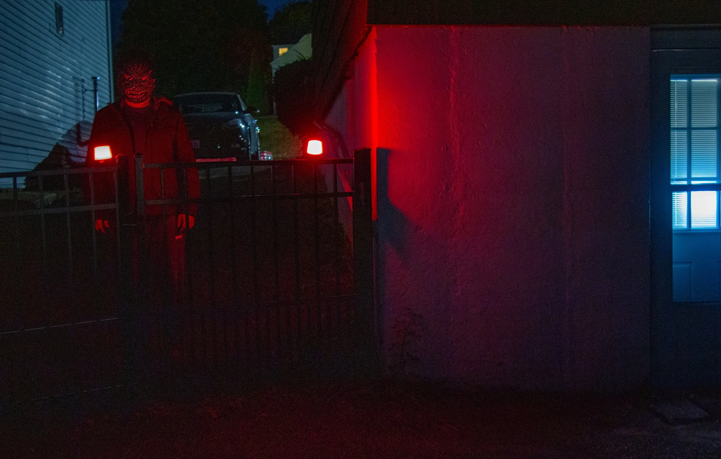 A person standing behind a gate illuminated by red lights at night, with a house on the right and a dark vehicle in the background.