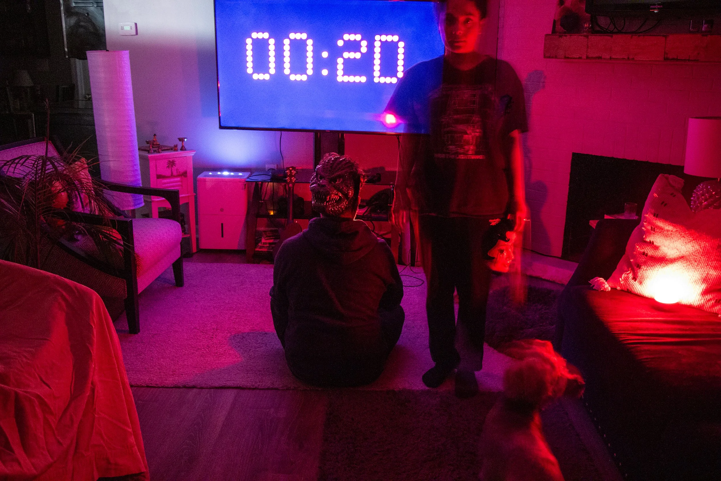 Living room decorated with pink and purple lighting, featuring a large digital timer on a TV screen showing 00:20. Two people are in the room: one standing, holding a dog, and the other sitting on the floor wearing a mask with a monster face. A dog i