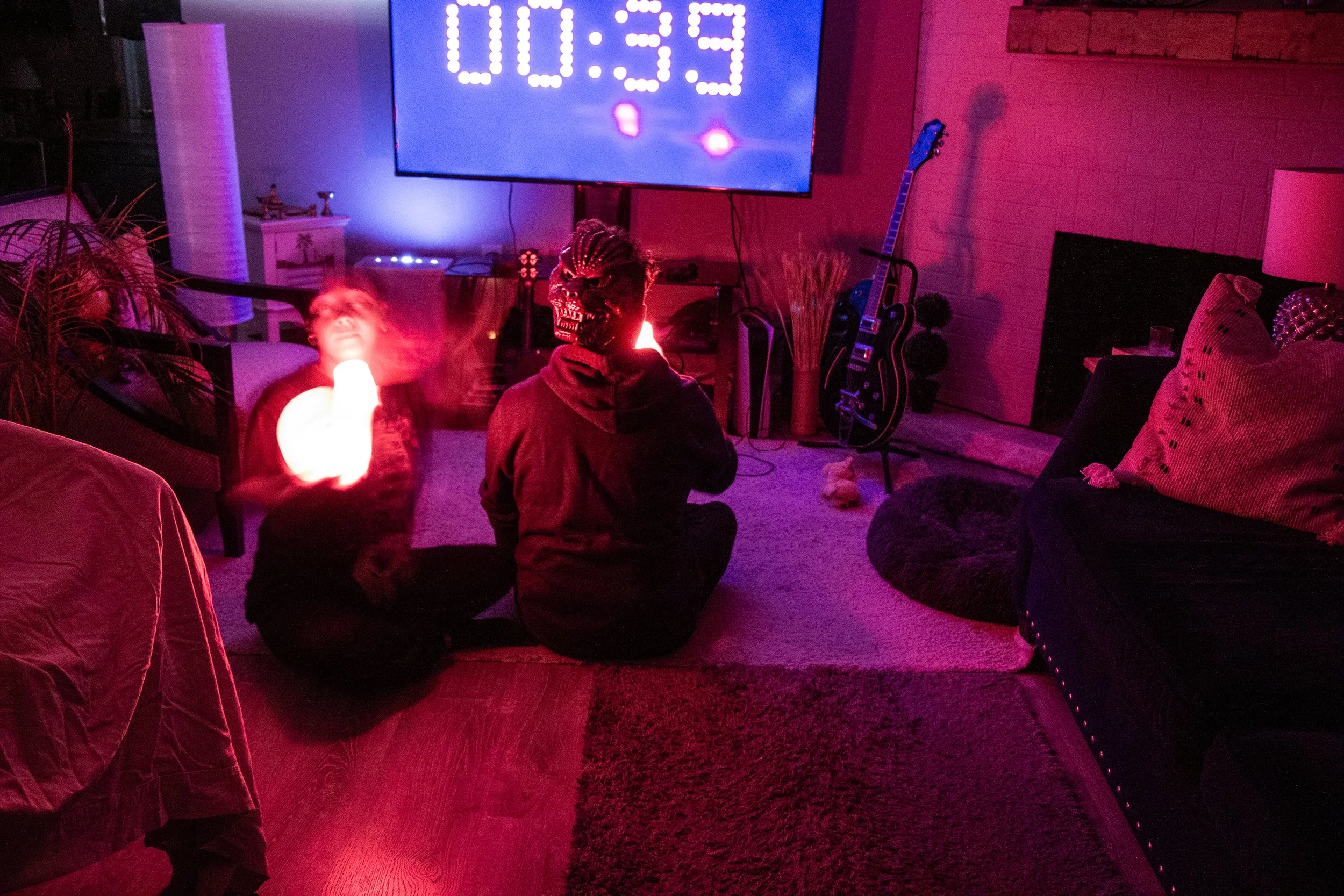 Two people sitting on the floor of a living room watching a large screen with a timer, surrounded by a guitar, houseplants, and pillows, in a room lit with red and purple lighting, with one person wearing a skull mask.