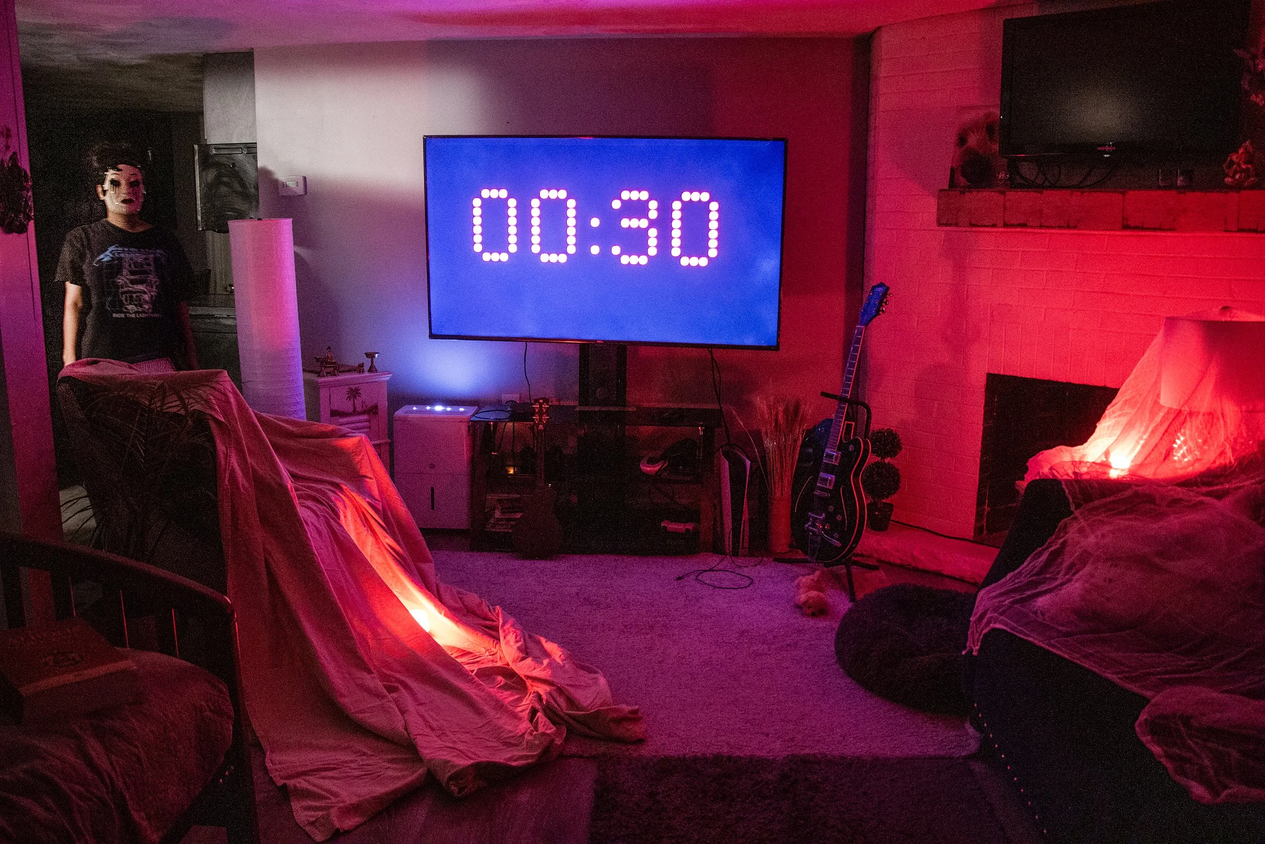 Living room with seating covered in sheets, a large TV displaying a countdown of 30 seconds, a guitar on a stand, and a person wearing a mask standing in the doorway. The ambient lighting is red and purple.