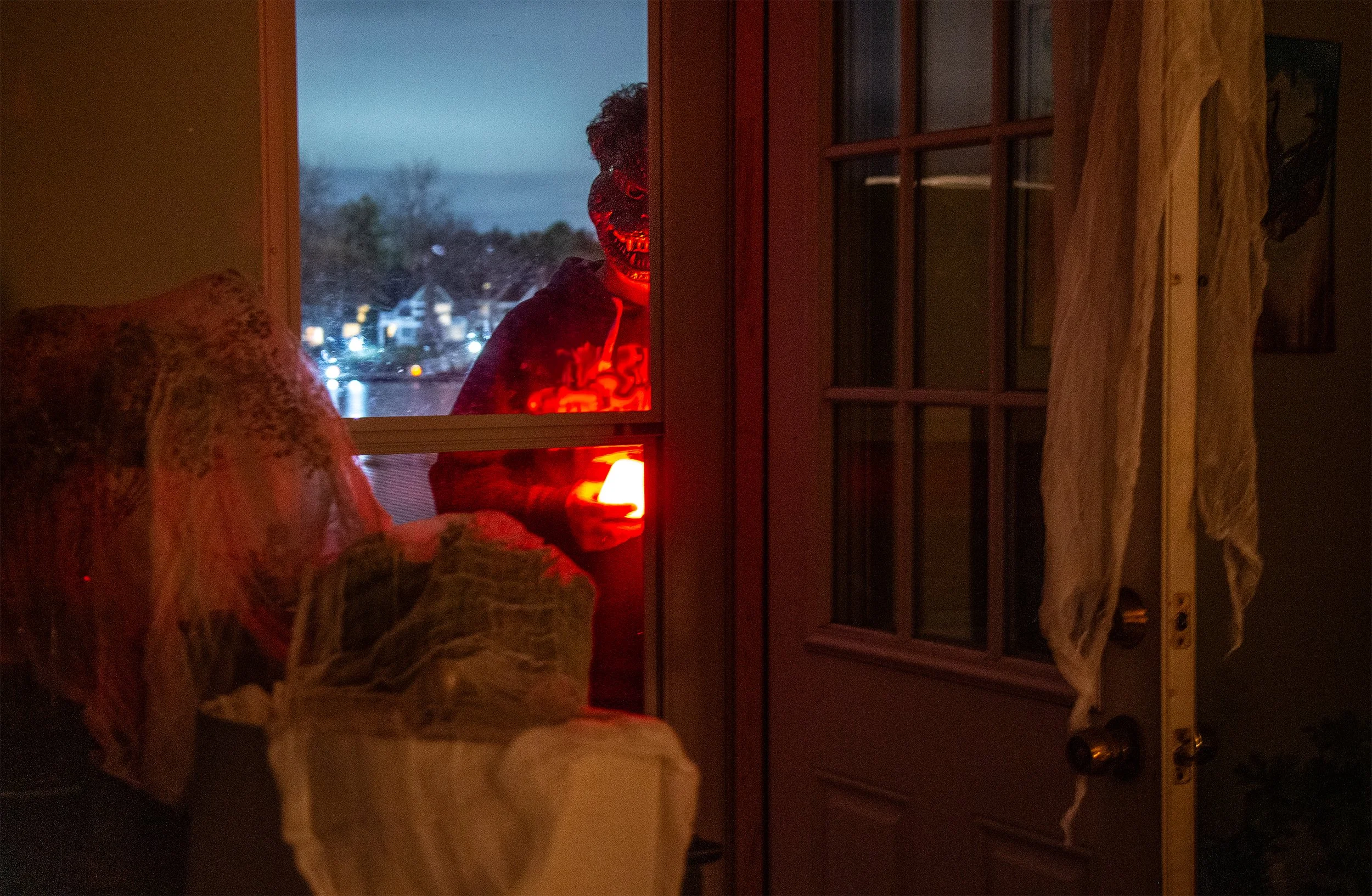 A person wearing a hooded sweatshirt with a glowing red skull mask is seen outside through a window at dusk, holding a lit candle inside.