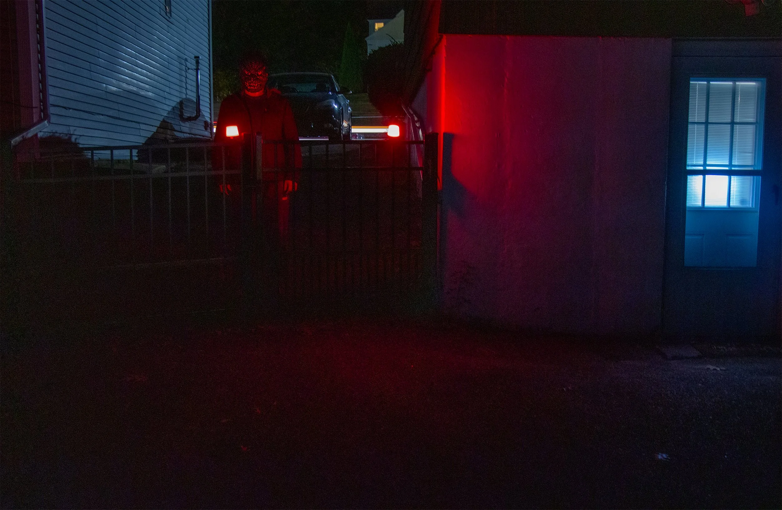 Person in dark clothing and a mask standing behind a metal gate, illuminated by red and blue lights at night.