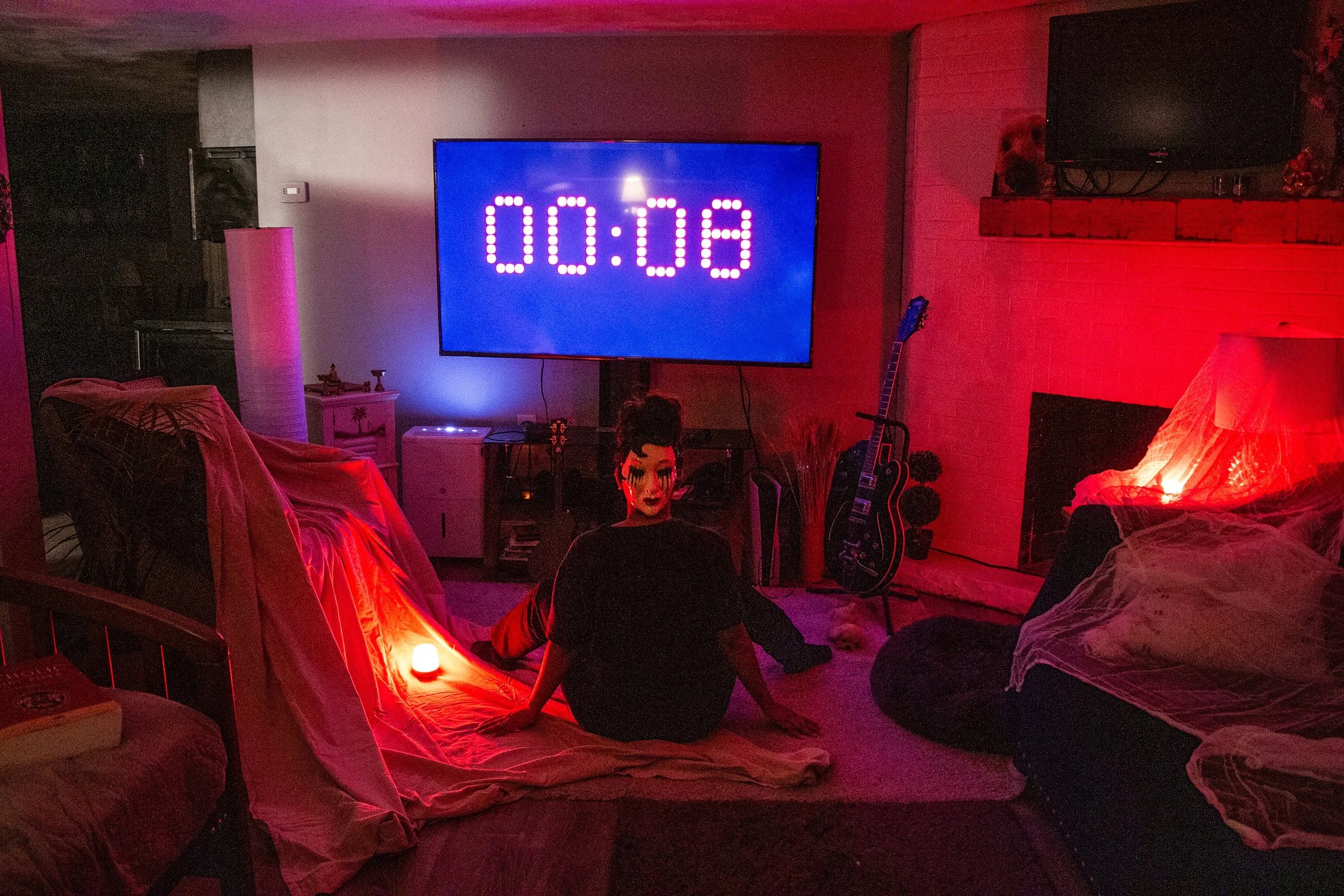A person with dark hair and makeup sitting on the floor in a dimly lit living room with pink and red lighting, a large digital timer on the wall showing 00:08, and various musical instruments including an electric guitar.