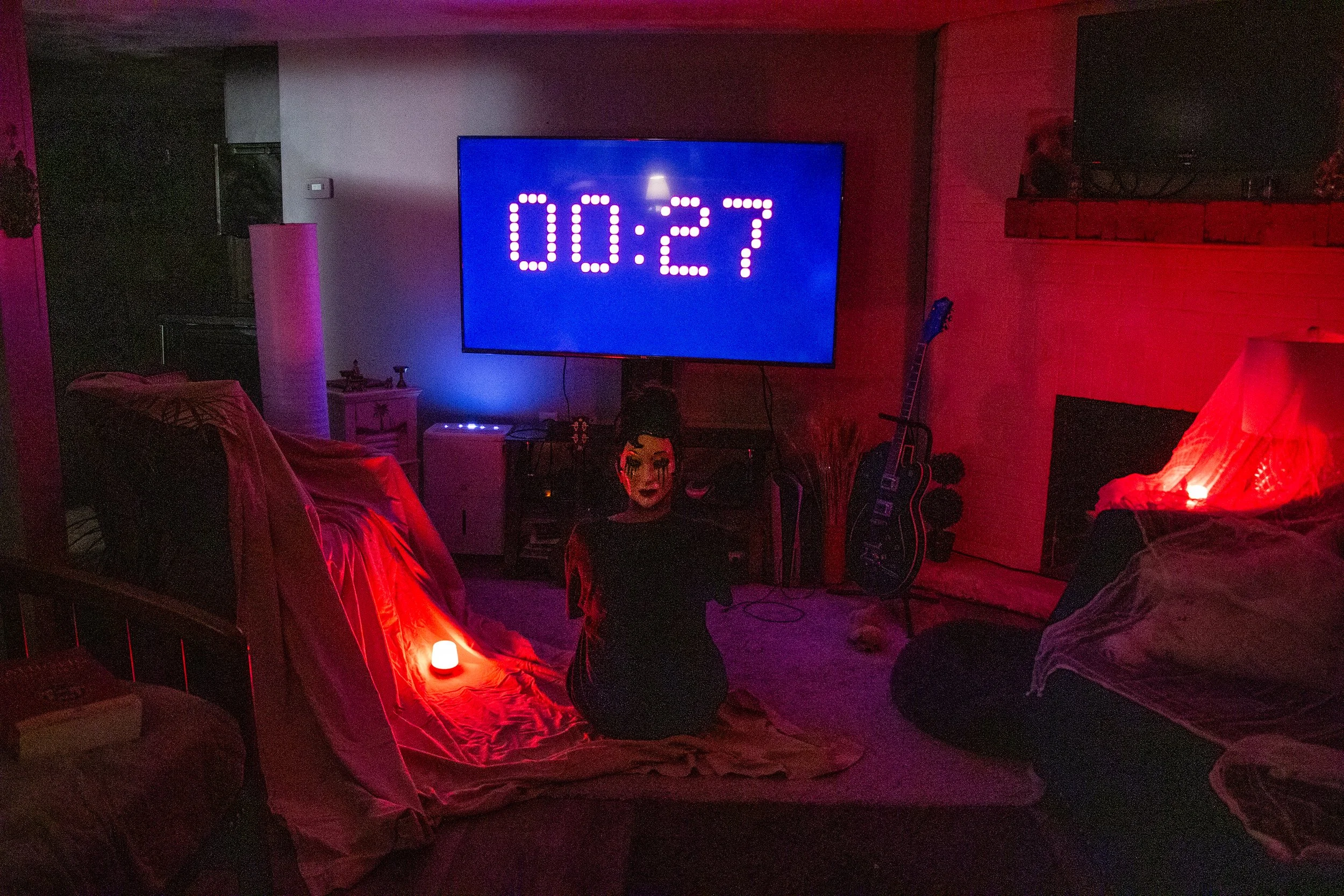 A dimly lit living room with red and purple ambient lighting. There is a large TV screen showing a countdown timer at 00:27. A woman dressed in black with dramatic makeup is sitting on the floor in front of the TV. To the left, there is a couch cover