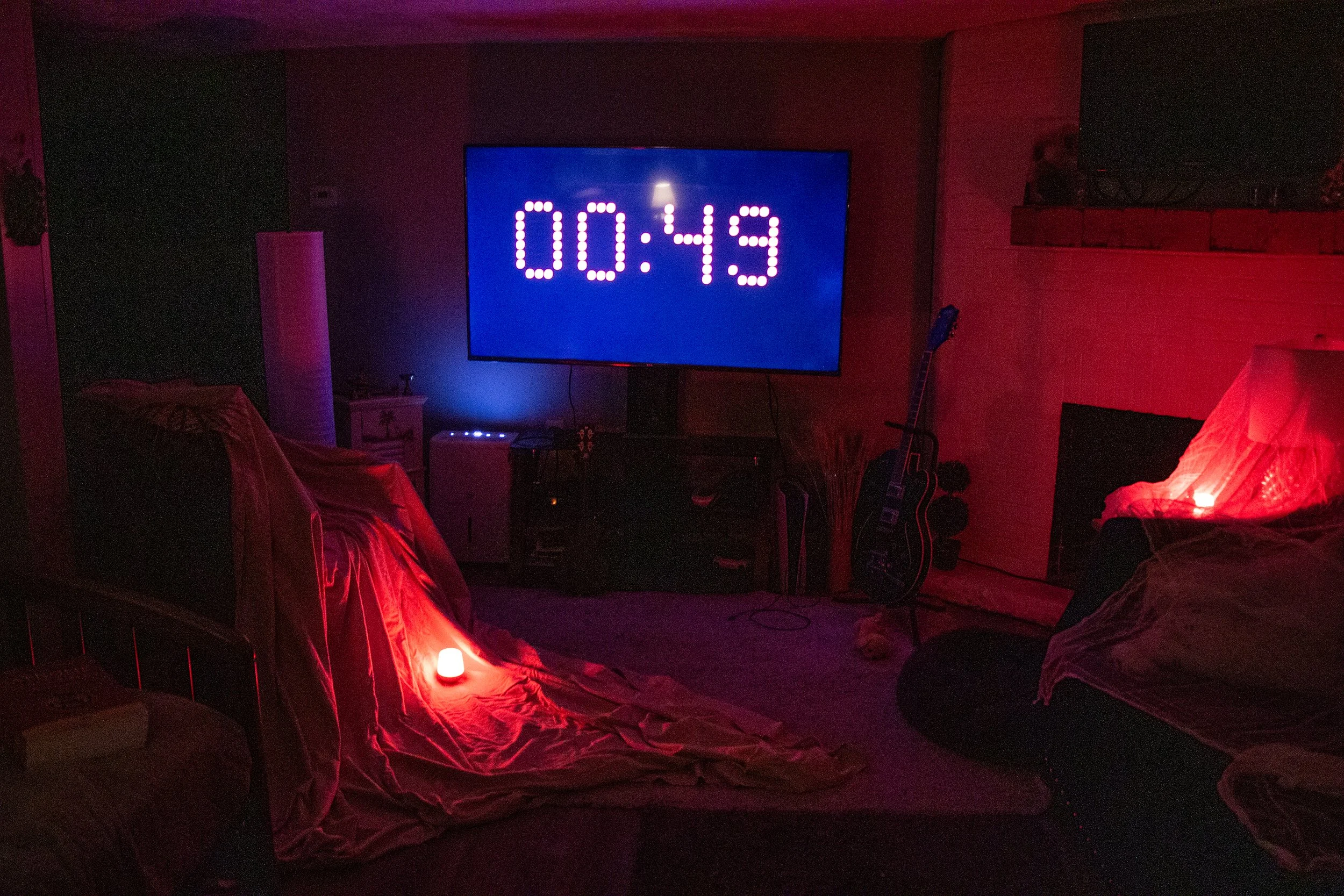 A dimly lit room with a large television displaying 00:43, two couches draped with cloth, and a glowing red light on a side table. A guitar is leaning against the wall near the fireplace.