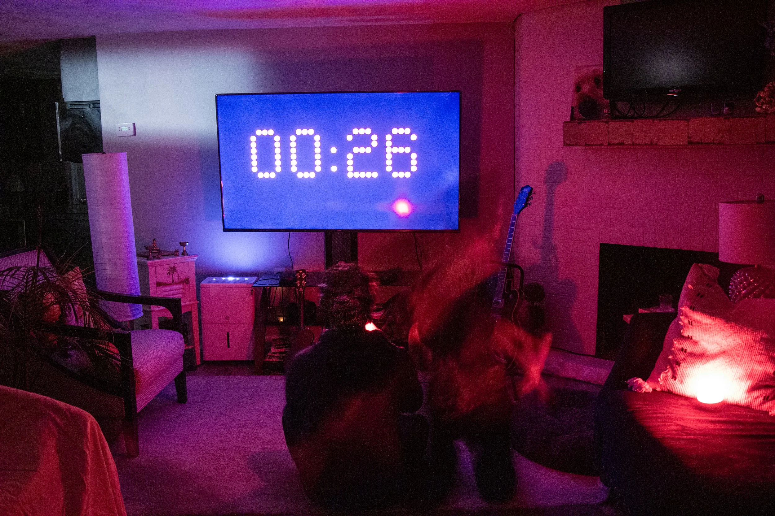 People watching a countdown on a large TV screen in a dimly lit living room with pink and blue ambient lighting.
