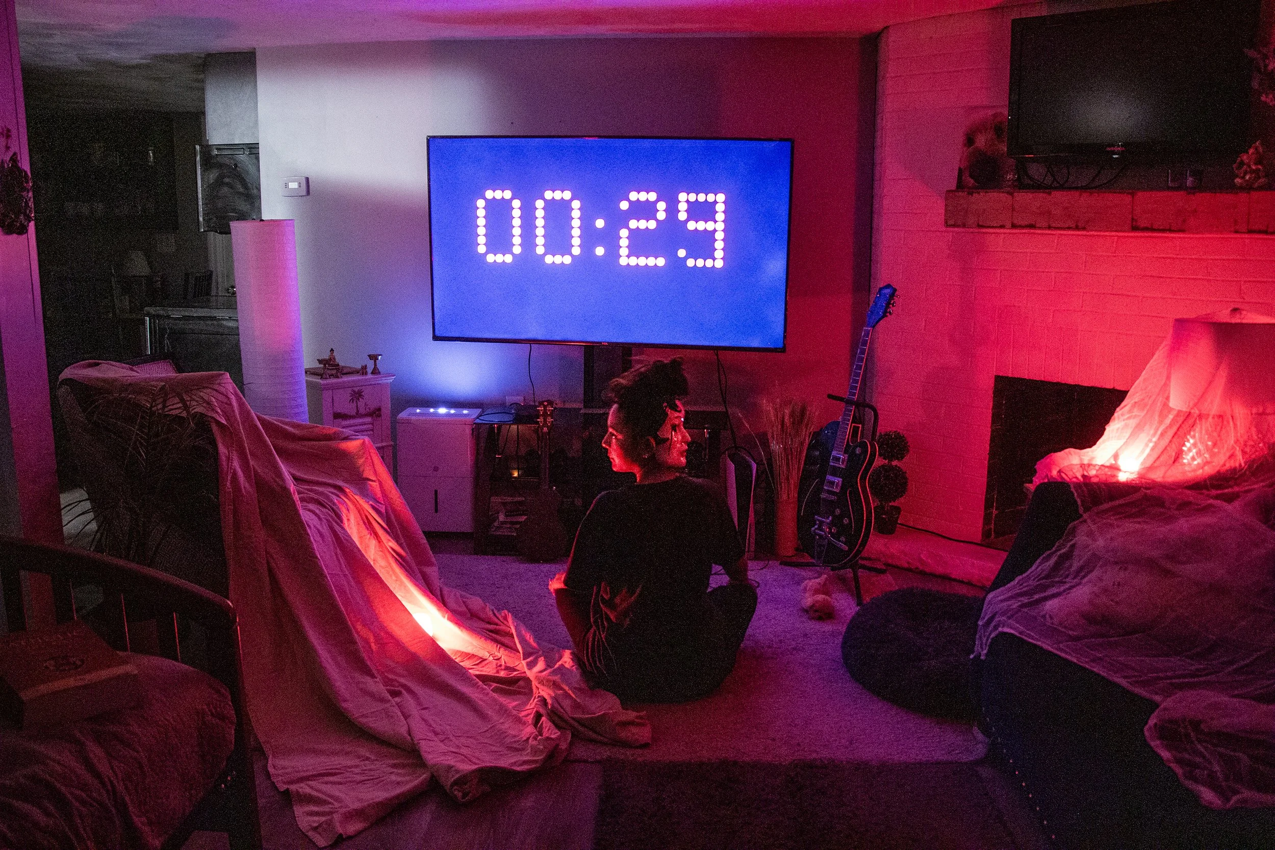 Living room with TV displaying a countdown timer, a woman sitting on the carpet, guitar leaning against the wall, and colorful lighting.
