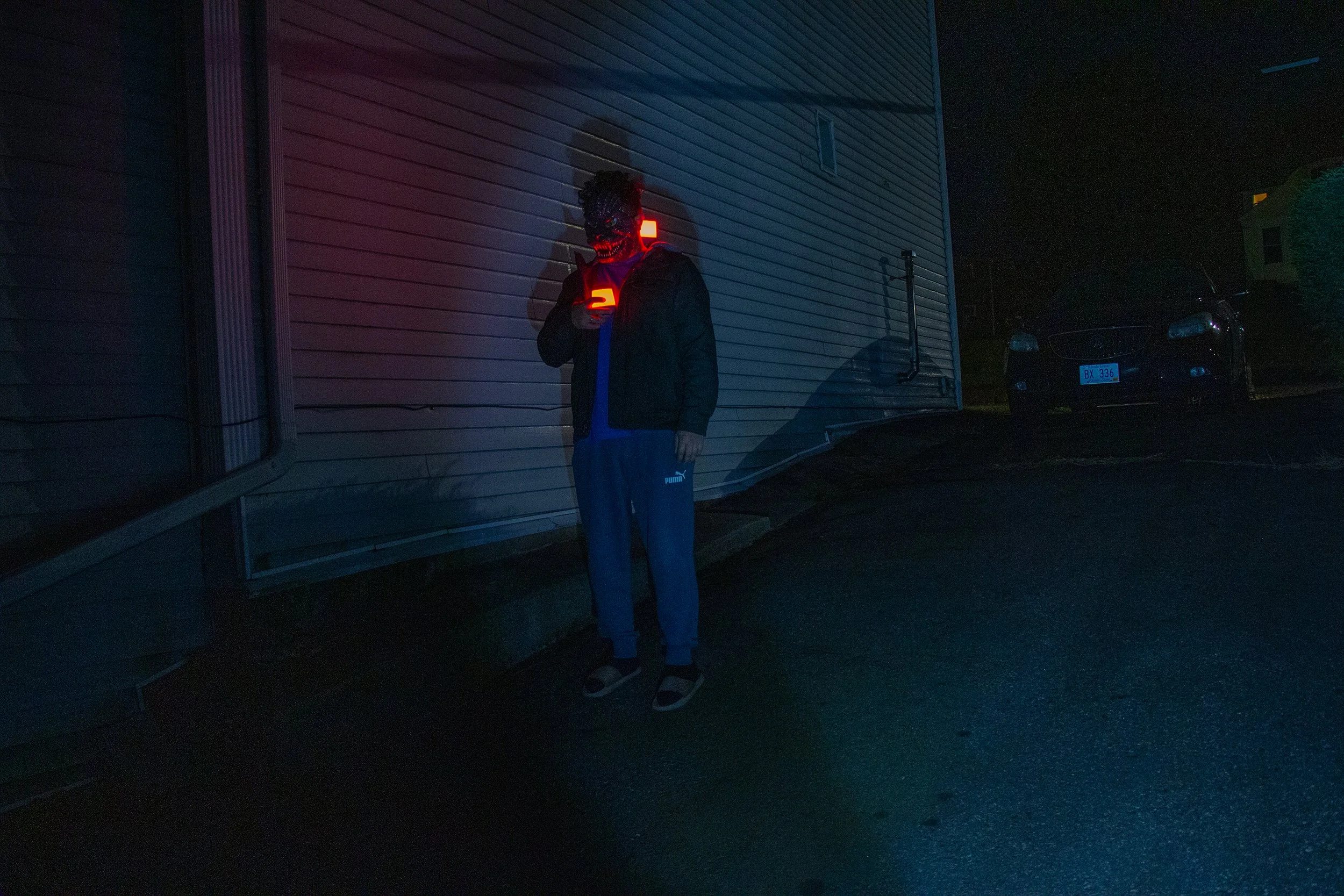 A person wearing a mask with a scary face design, a black jacket, blue pants, and sandals standing against a building at night, holding a glowing object in their hand.