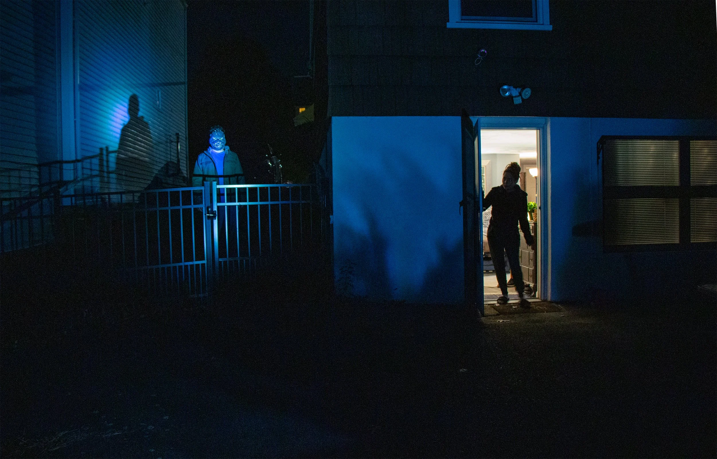 Night scene with three people near a house: one walking out of the house, two standing outside on the left side, illuminated by blue light, with curtains and interior lights visible inside.