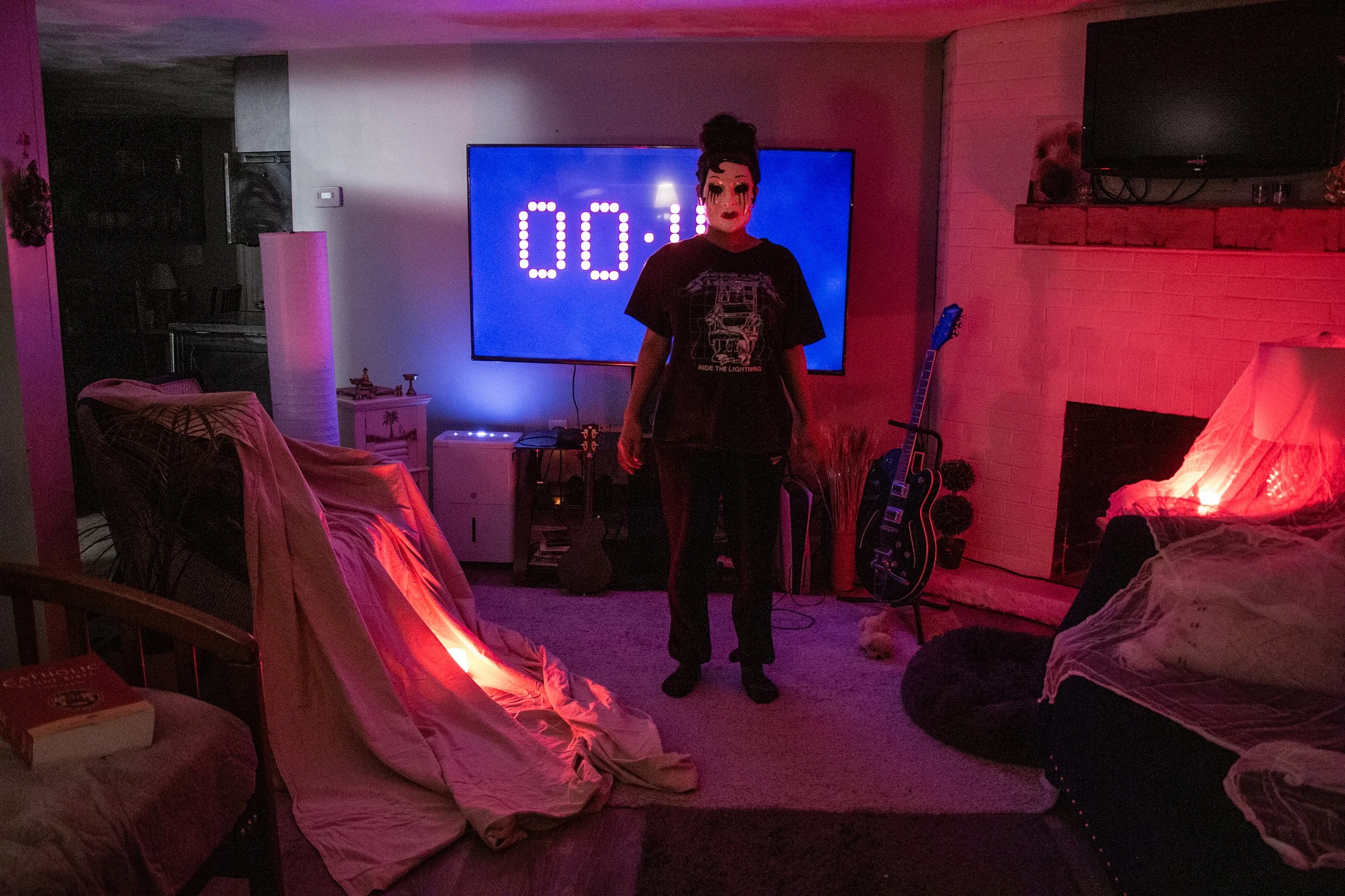 A person wearing a mask and black clothing standing in a dimly lit living room with red and blue lighting. A large TV with an illuminated timer is on the wall behind them, and various furniture and decorations are visible around the room.