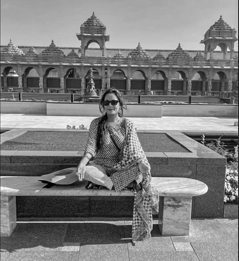 Person sitting on a marble bench in front of a traditional architectural building with ornate columns and domes, wearing sunglasses and holding a hat.