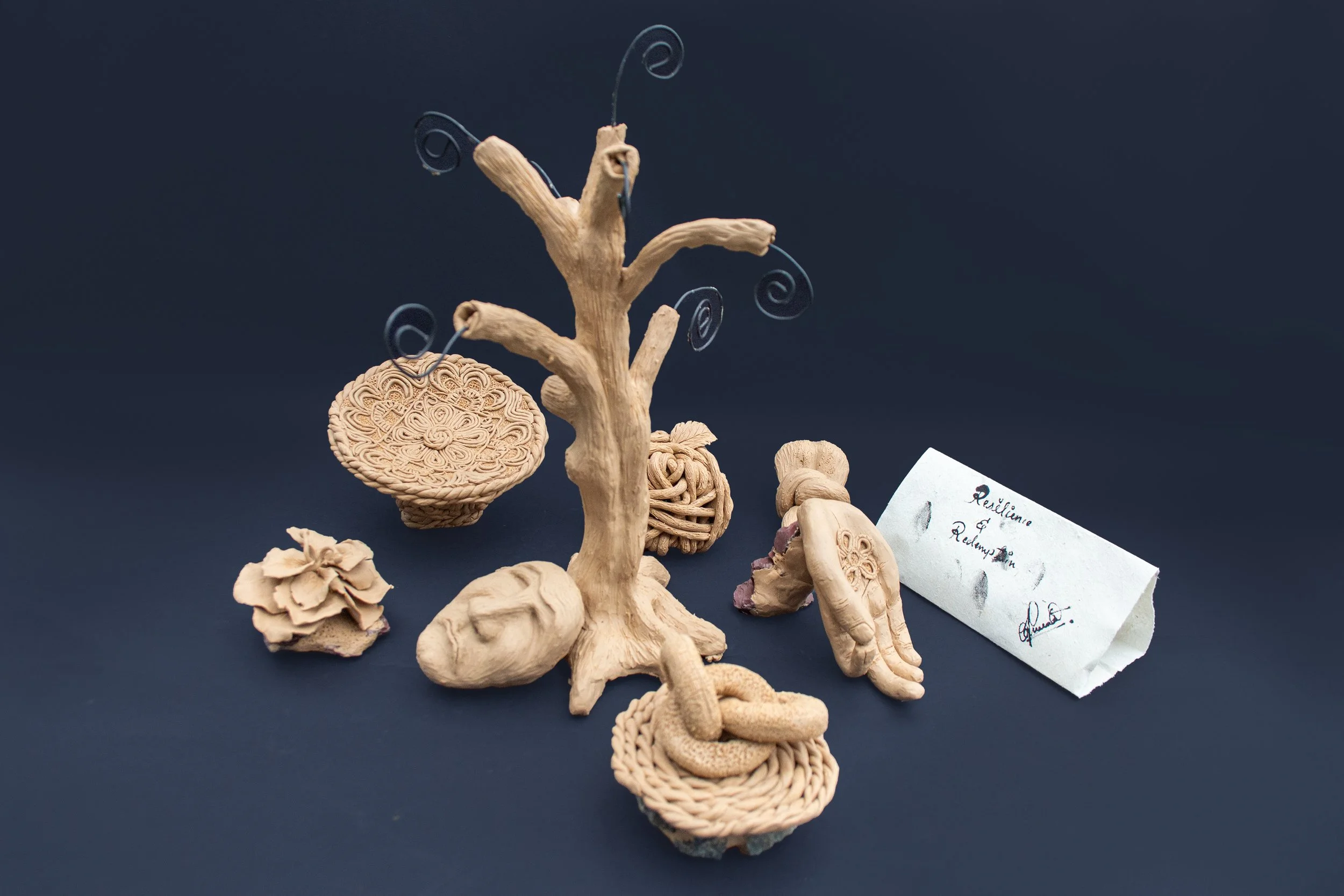 Collection of beige ceramic sculptures resembling a tree, a frog, a flower, a snake, and decorative objects, arranged on a dark surface with a handwritten note reading 'Resilience & Rejuvenation'.