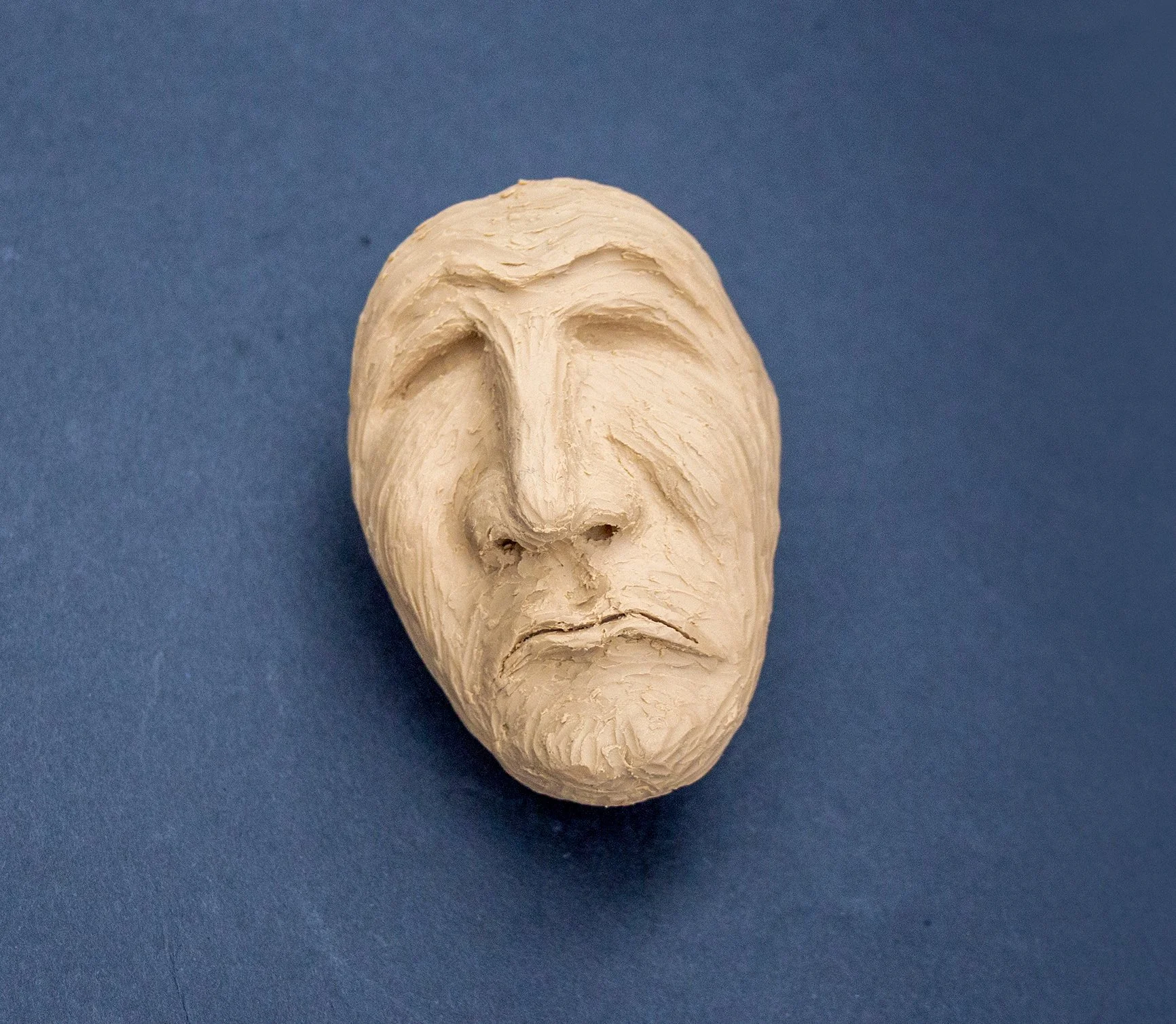 Sculpted clay face with a large nose, closed eyes, and a sad expression, against a blue background.
