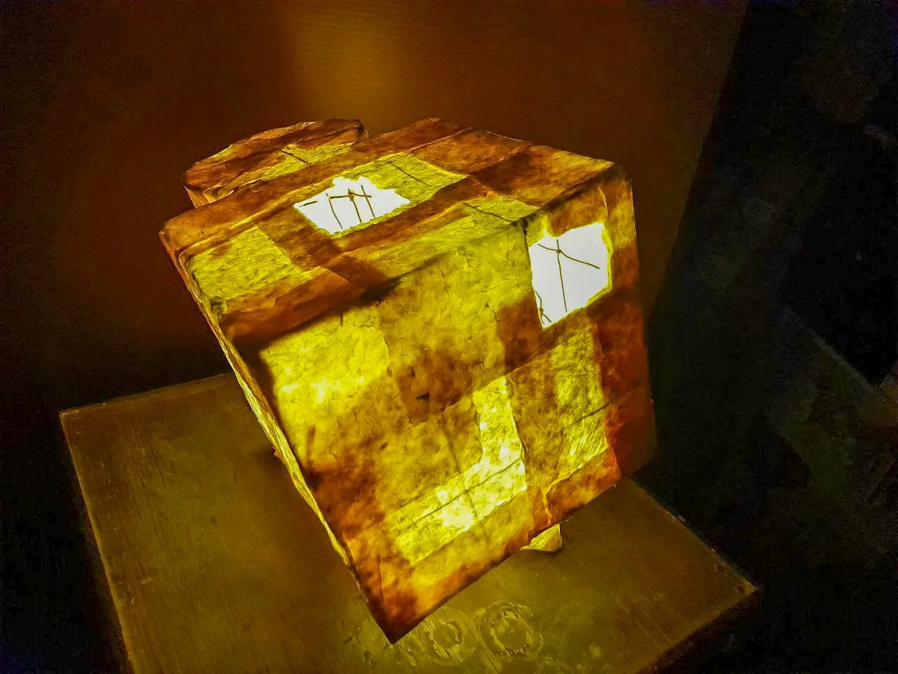 Illuminated cube lamp with stained glass panels, placed on a wooden surface in a dimly lit room.