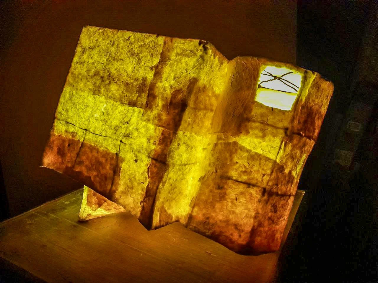Illuminated, abstract sculpture made of yellow and orange translucent material, resembling a geometric, folded shape with a small cutout window, placed on a wooden surface in a dark room.