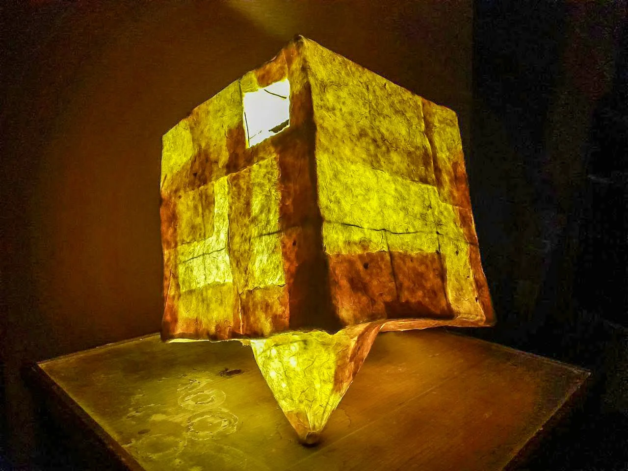 Illuminated paper lantern with a geometric, cube-like shape, glowing in yellow and orange hues, resting on a wooden surface