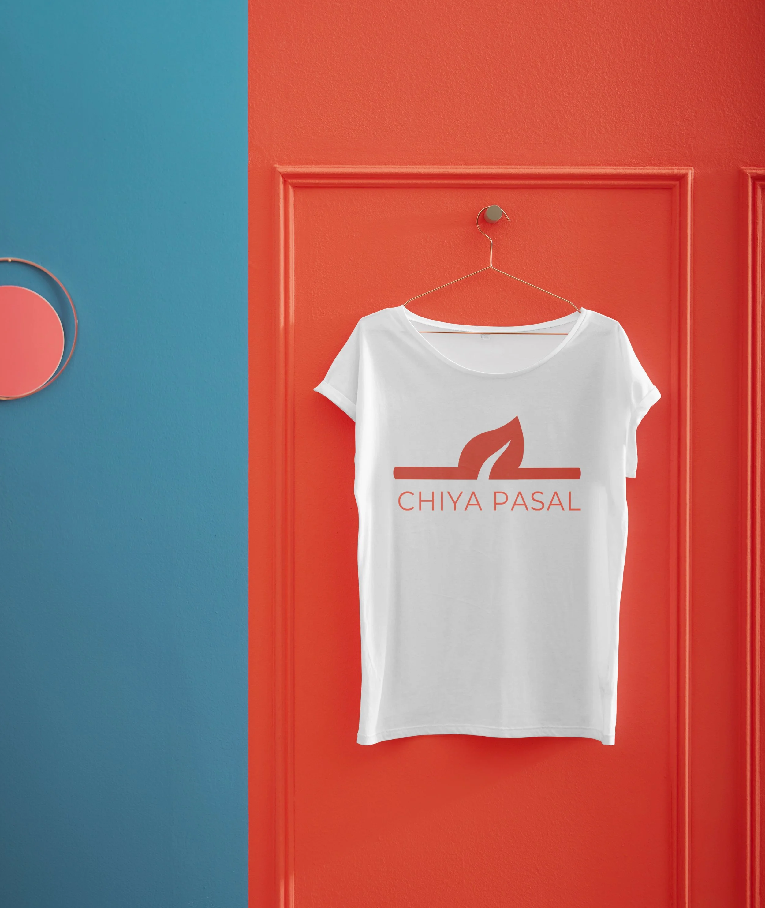 White T-shirt hanging on a hook with a leaf logo and the words 'Chiya Pasal' printed in red, against a red and blue wall background.