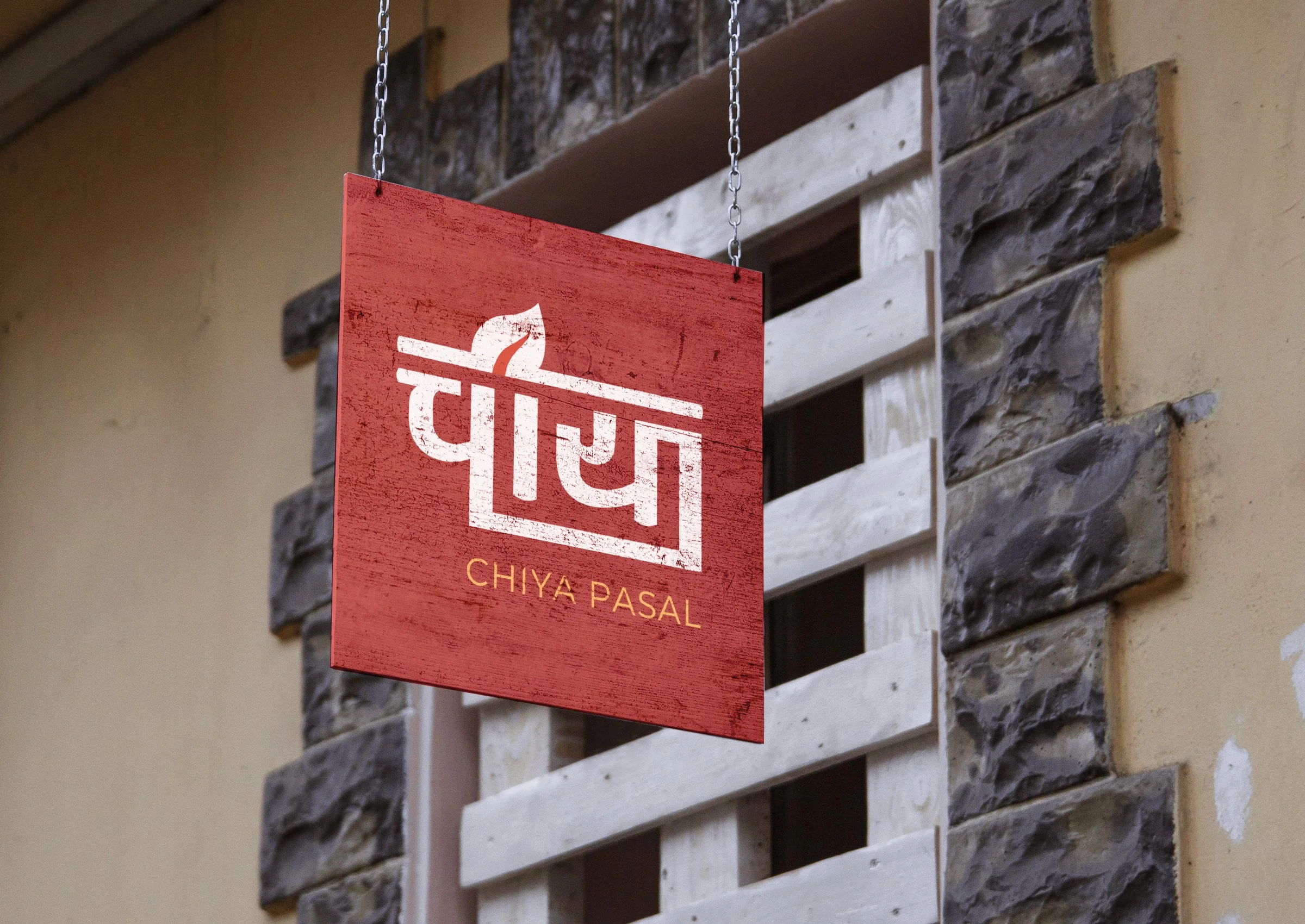 A red sign with white stylized Hindi script and the words "Chiya Pasal" hanging on a chain in front of a stone and wooden wall.