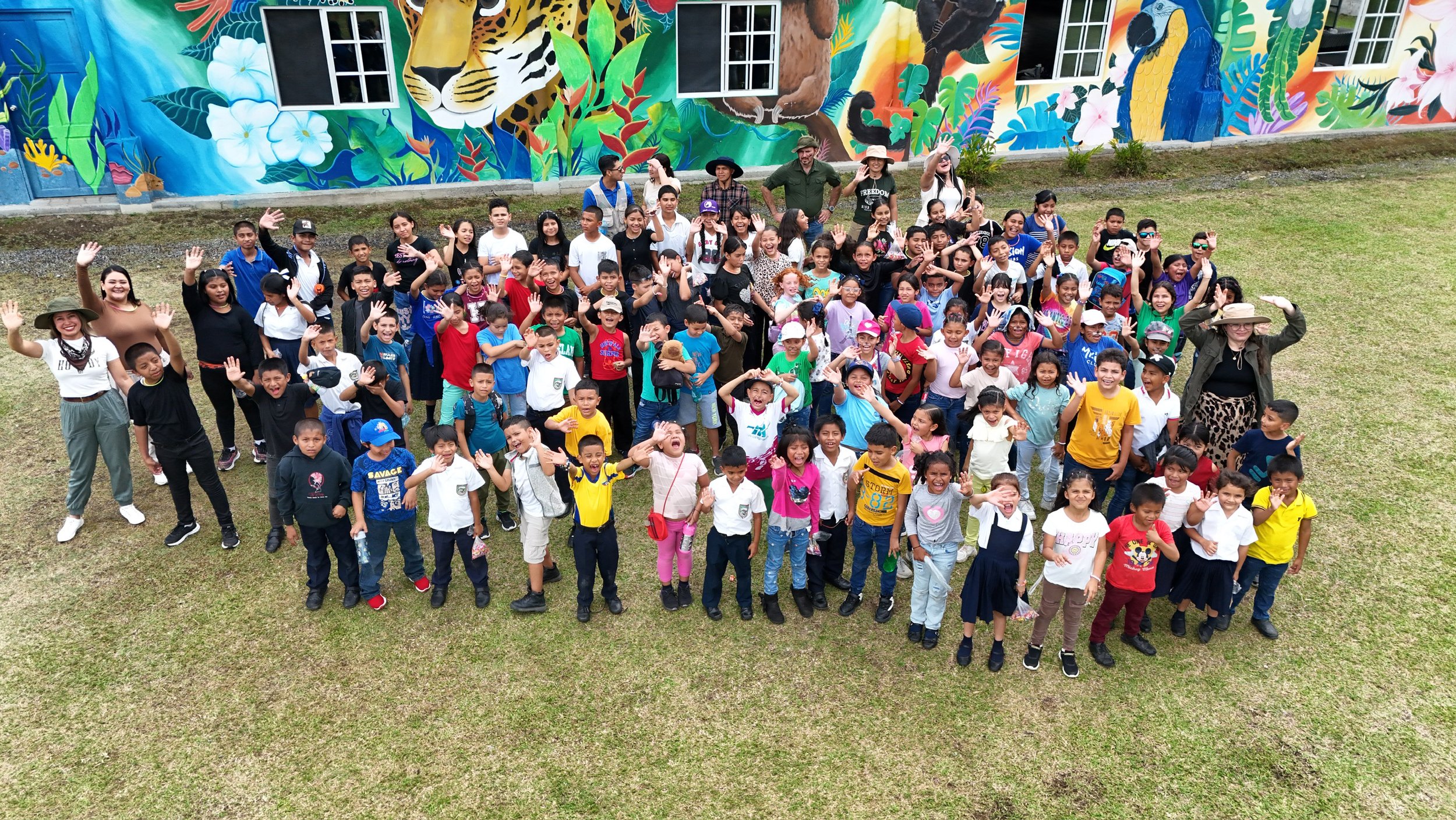 A large group of children and some adults smiling and waving, gathered outdoors on grass in front of a colorful mural depicting jungle animals and plants.