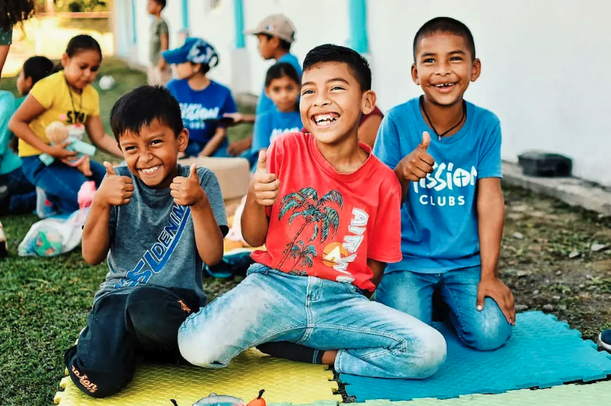 Three smiling children sitting on mats outdoors, giving thumbs up. More children in the background, some standing and some sitting, with a building and grass visible.