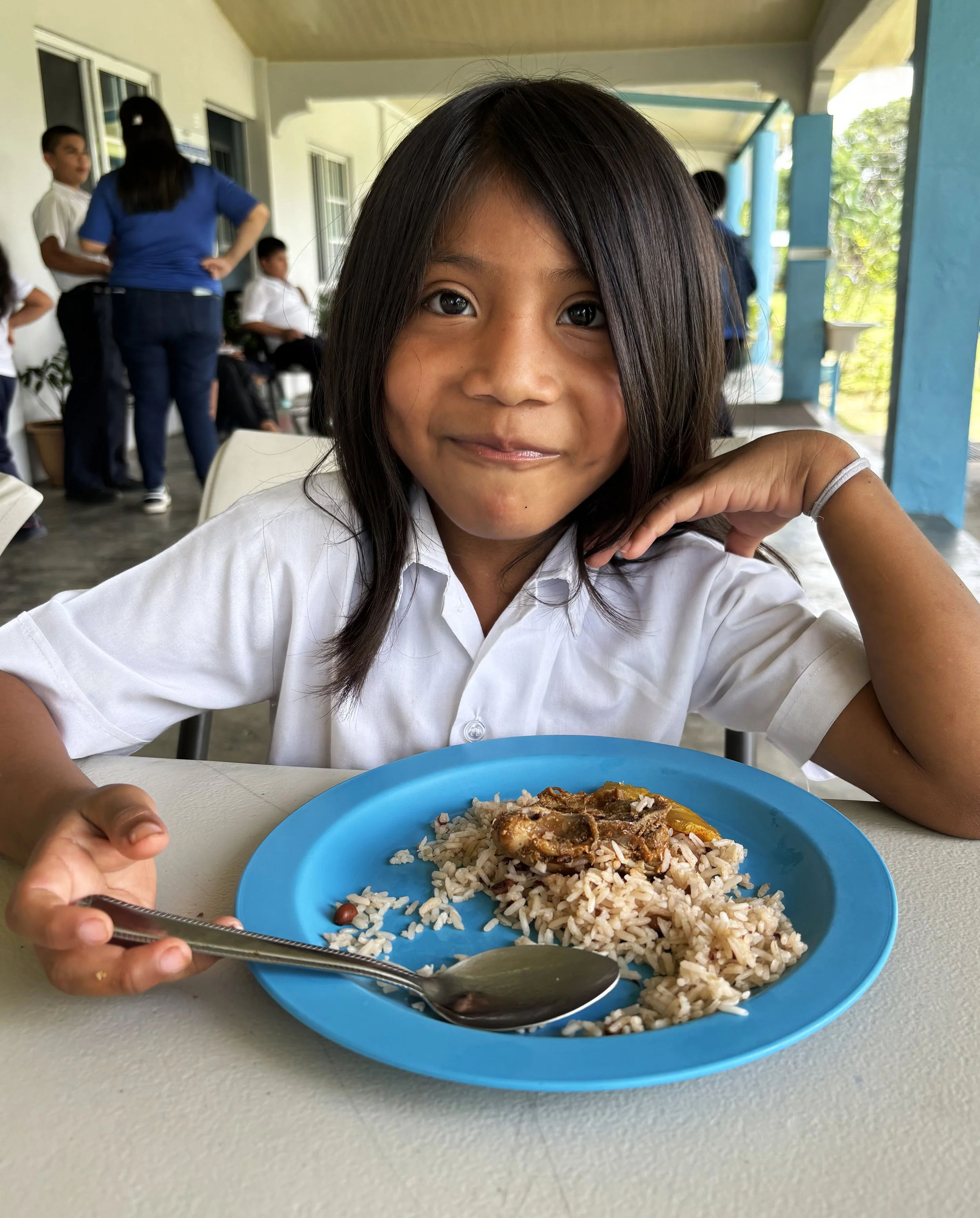 A young girl with long dark hair and a white shirt sits at a table with a plate of rice and meat, smiling at the camera. In the background, other children and adults are standing and sitting in a school or cafeteria setting.
