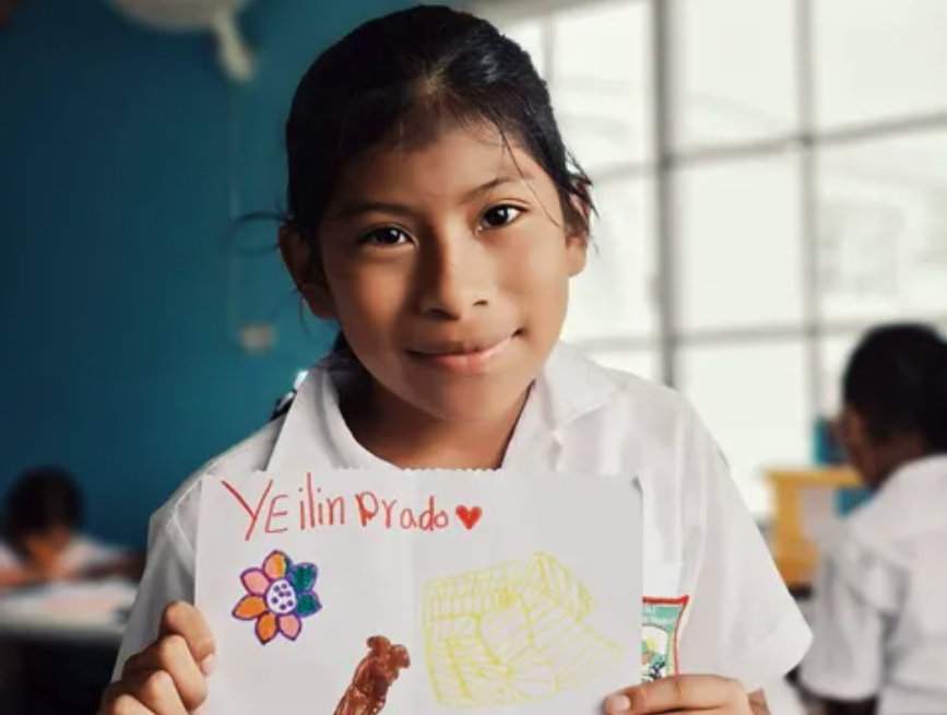 Young girl with dark hair in a ponytail holding a drawing sheet with her name and pictures, inside a classroom with other students and large windows in the background.